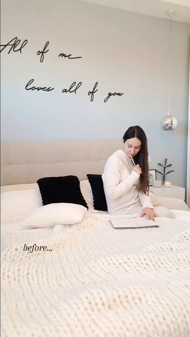 Woman in cream sweatsuit sits on bed with chunky knit blanket, writing in notebook. Wall art reads "All of me loves all of you."