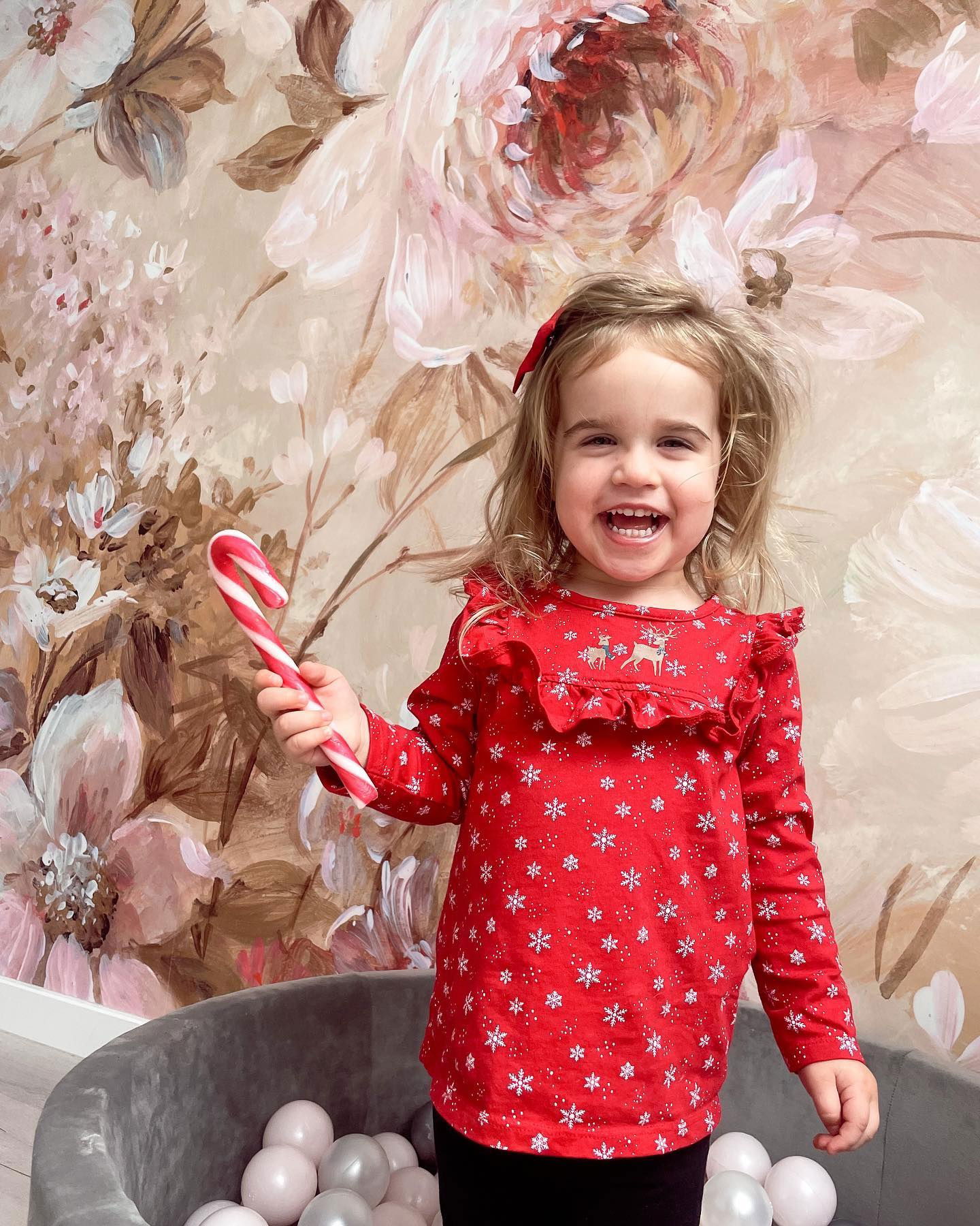 Lächelnder Kleinkind in rotem Schneeflockenhemd hält Zuckerstange vor Blumentapete und Bällebad.