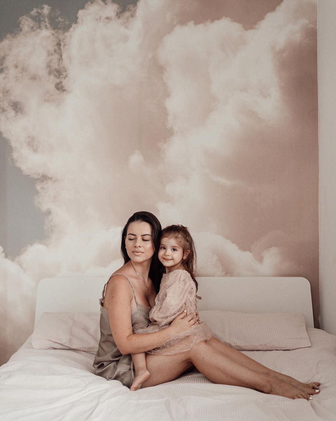 A woman in a slip dress holds a child in a pink dress on a bed.
