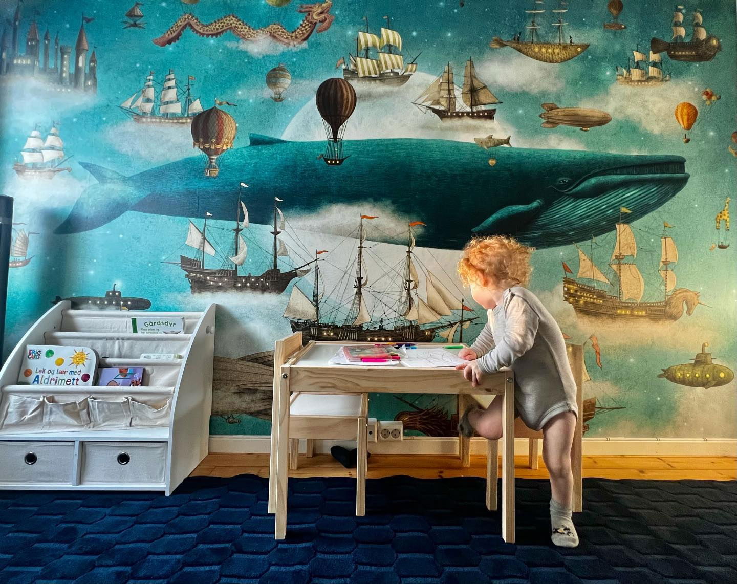 Child drawing at a table in a room with a whimsical whale and ship wallpaper.