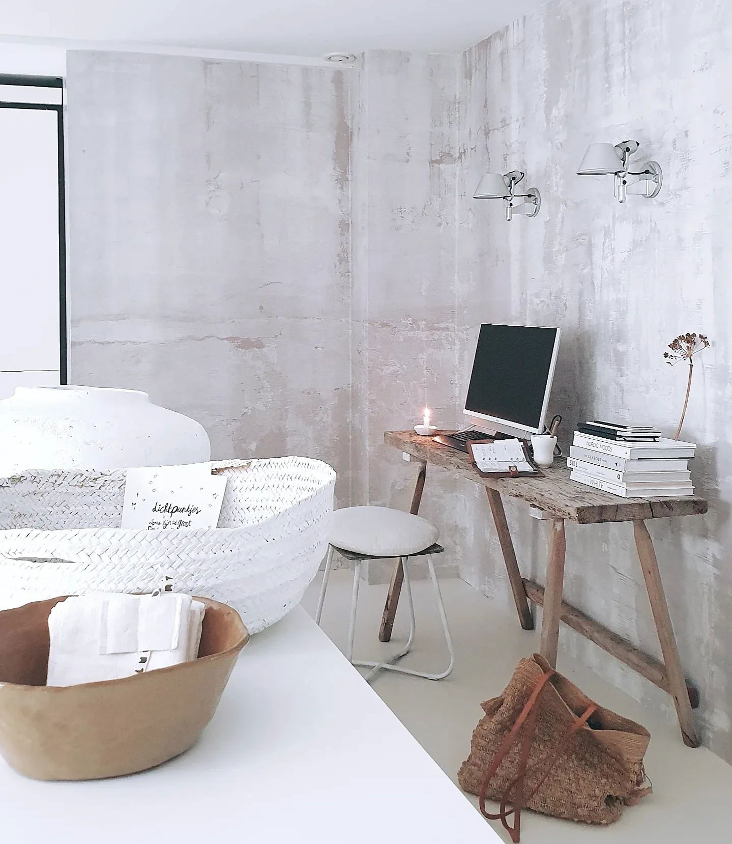 Minimalist workspace with computer on rustic wood desk, stool, and woven baskets against textured white wall.