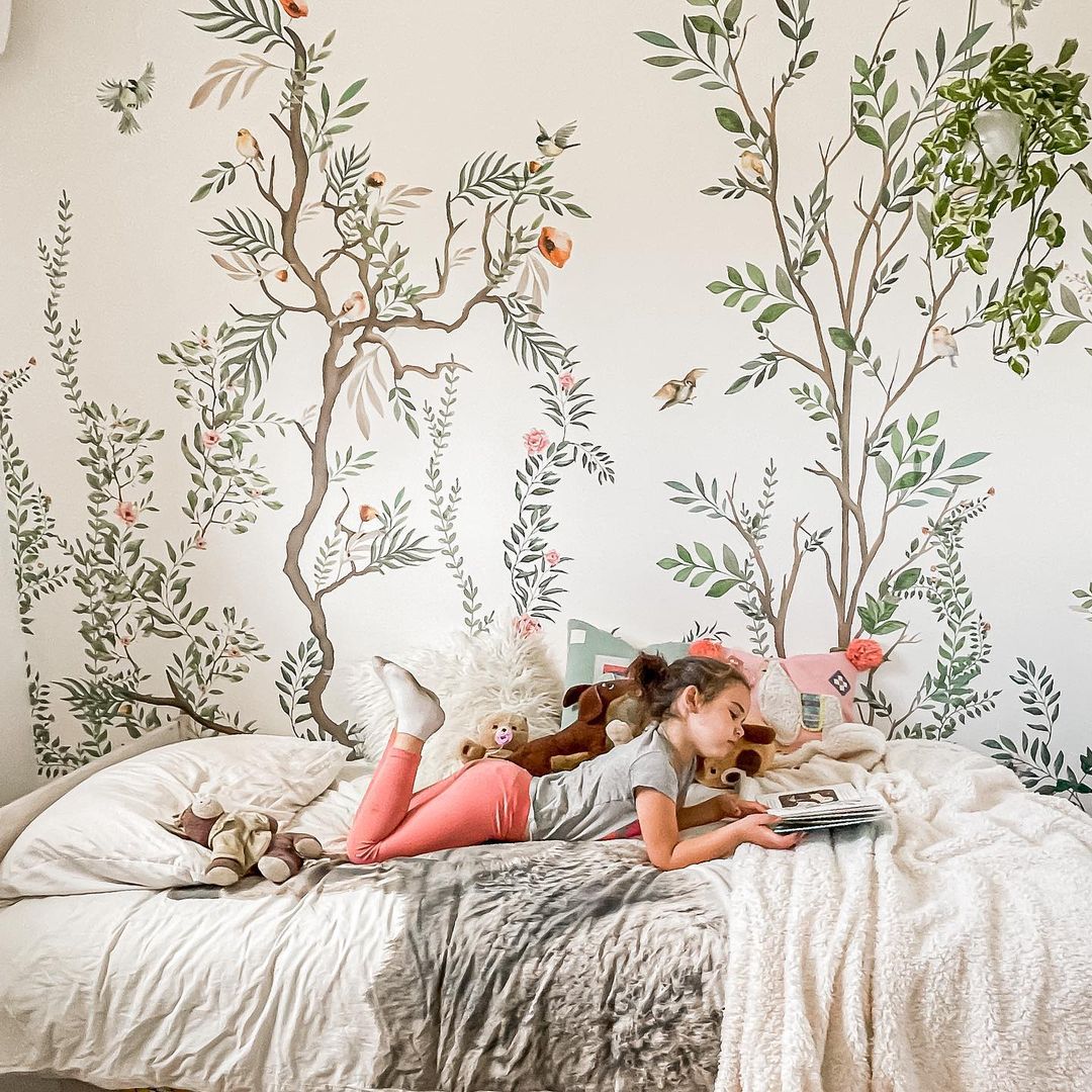 A girl reads on a bed with a floral mural. She wears pink leggings and a gray shirt, surrounded by stuffed animals.