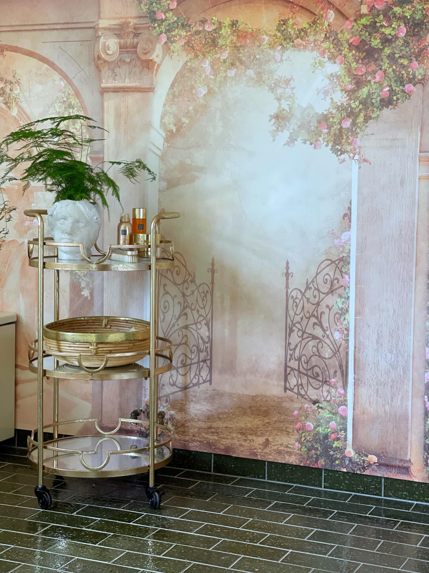 Gold bar cart with plant, bottles, and basket in front of a floral archway mural.