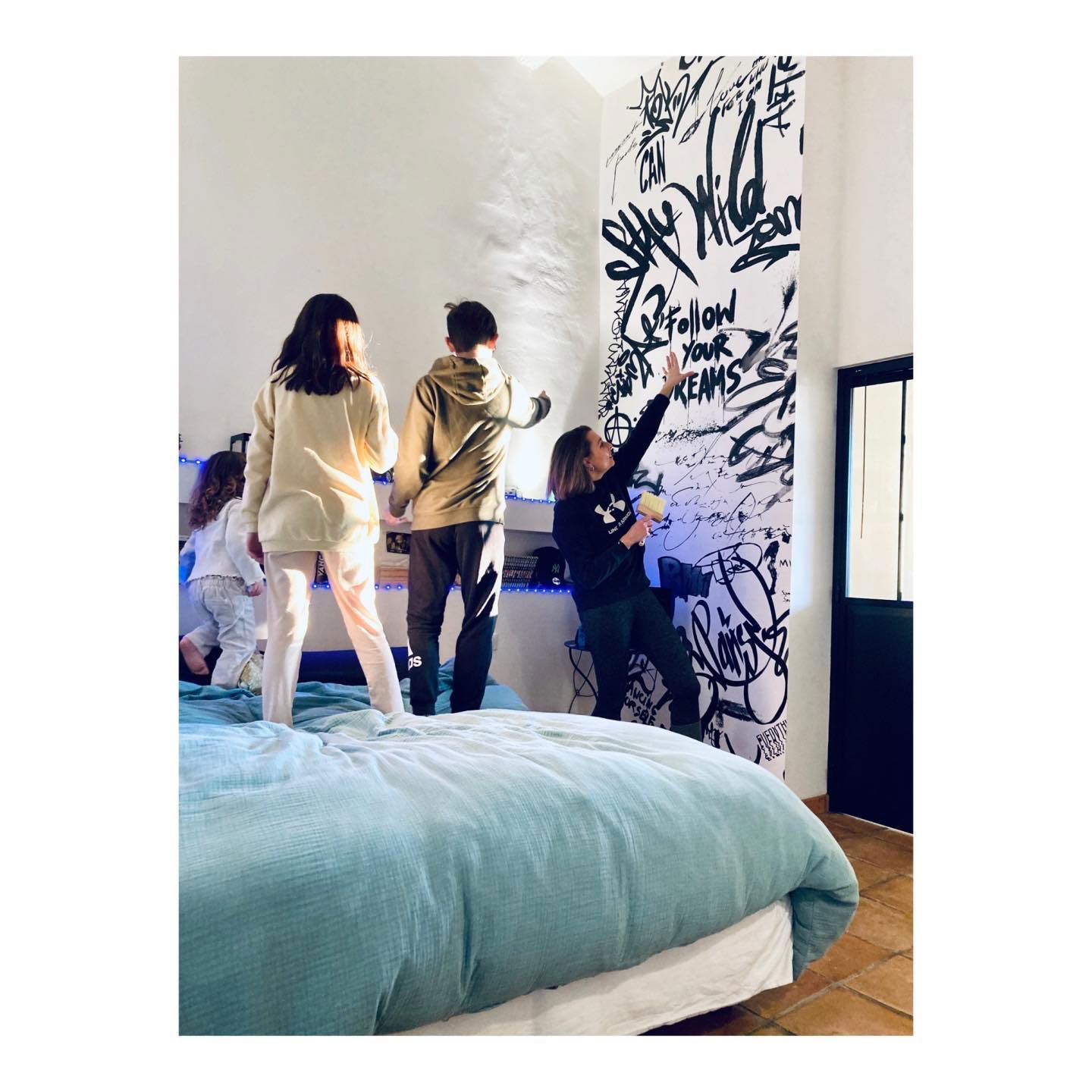A family stands on a bed in front of a wall mural with black graffiti.