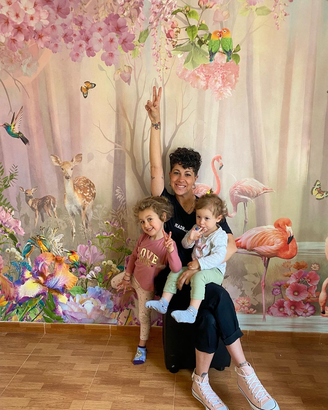 En kvinna poserar med två små barn framför en färgstark muralmålning med blommor och djur.