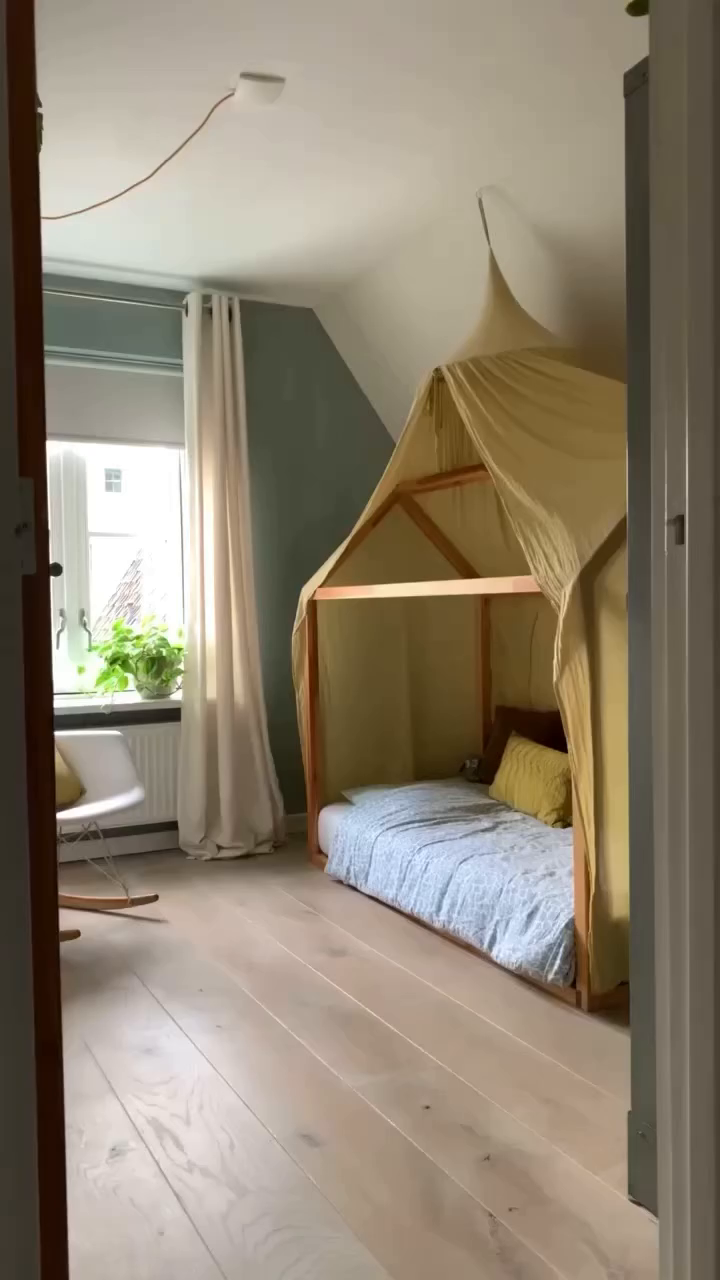 Bedroom with a wooden canopy bed draped in yellow fabric, light wood floors, and a white rocking chair.