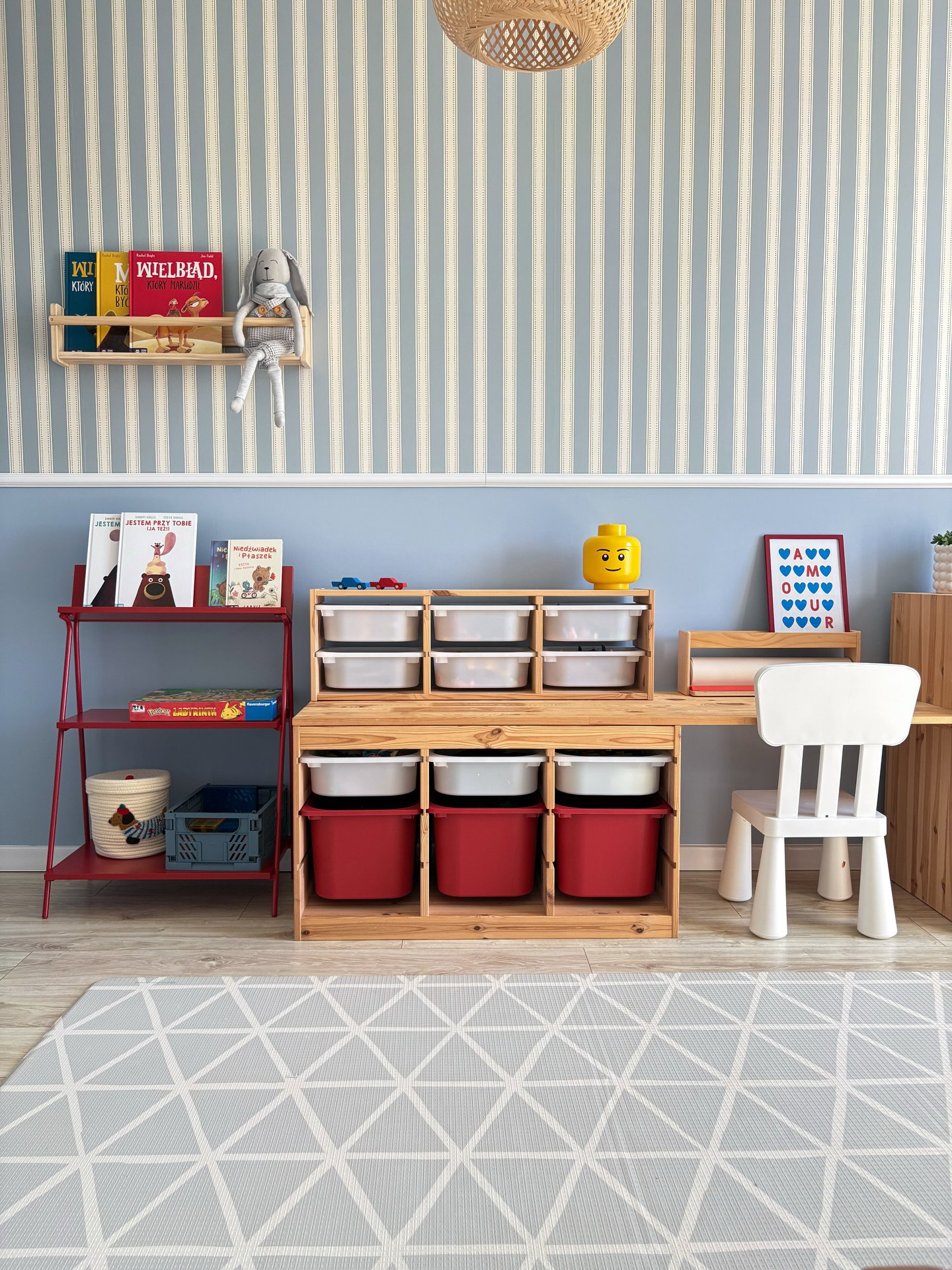 Ein Kinderspielzimmer mit blau-weiß gestreiften Wänden. Es verfügt über ein rotes Metallregal, eine Holzaufbewahrungseinheit mit roten und weißen Behältern, einen angeschlossenen Holztisch, einen weißen Kinderstuhl und einen grauen geometrischen Teppich.