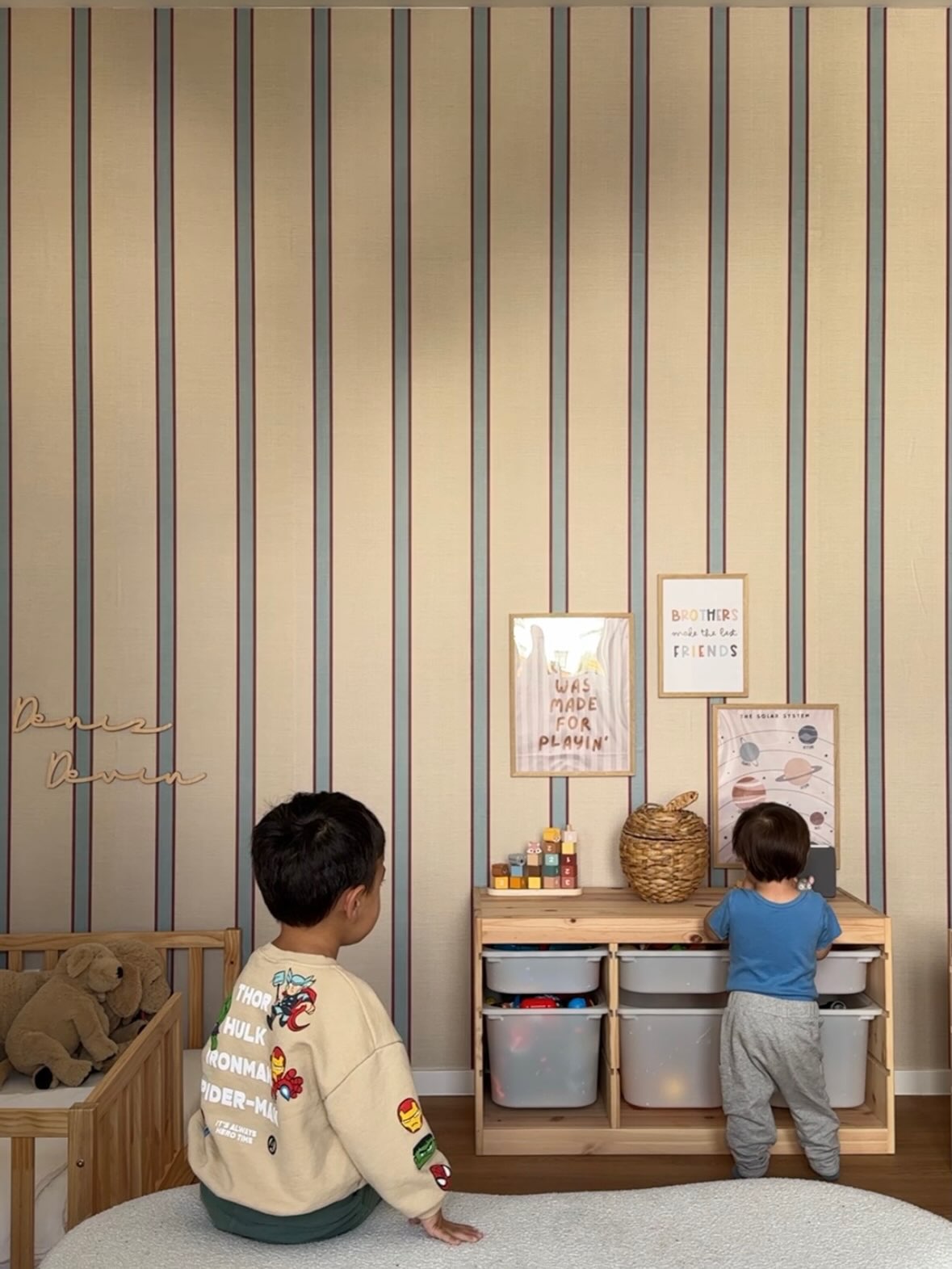 Dos niños pequeños en una sala de juegos. Un niño con una sudadera color beige con calcomanías de superhéroes se sienta en una alfombra, mirando a un niño más pequeño en una unidad de juguetes de madera. La pared tiene papel tapiz a rayas.