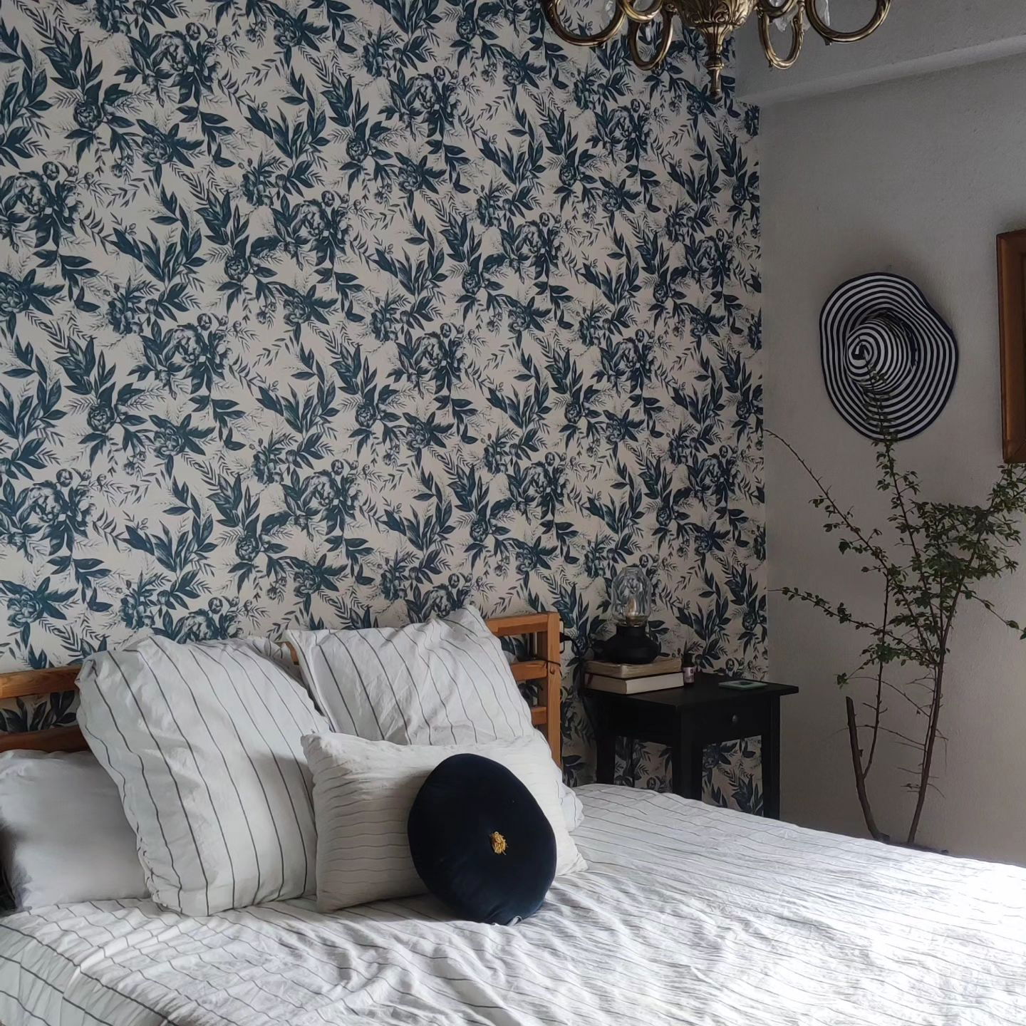 Soveværelse med blomstret tapet, stribet sengetøj og sort sengebord. En stribet hat hænger på væggen.