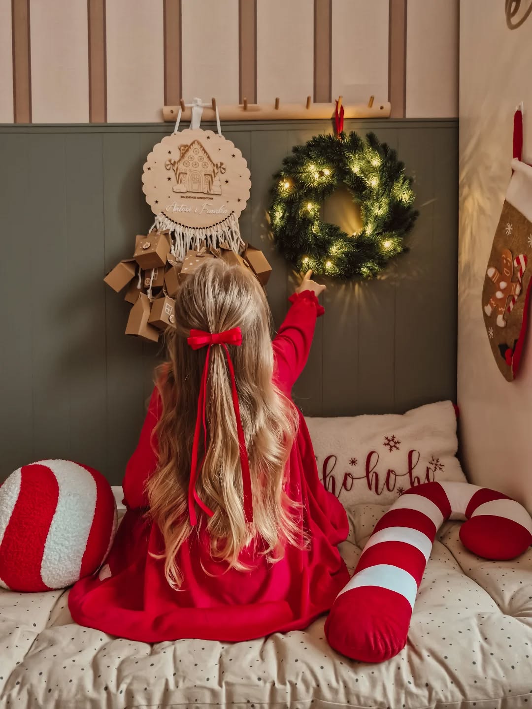 A blonde girl in a red dress reaches for a lit Christmas wreath on the wall. Christmas décor includes candy cane pillows, stocking, and advent calendar.