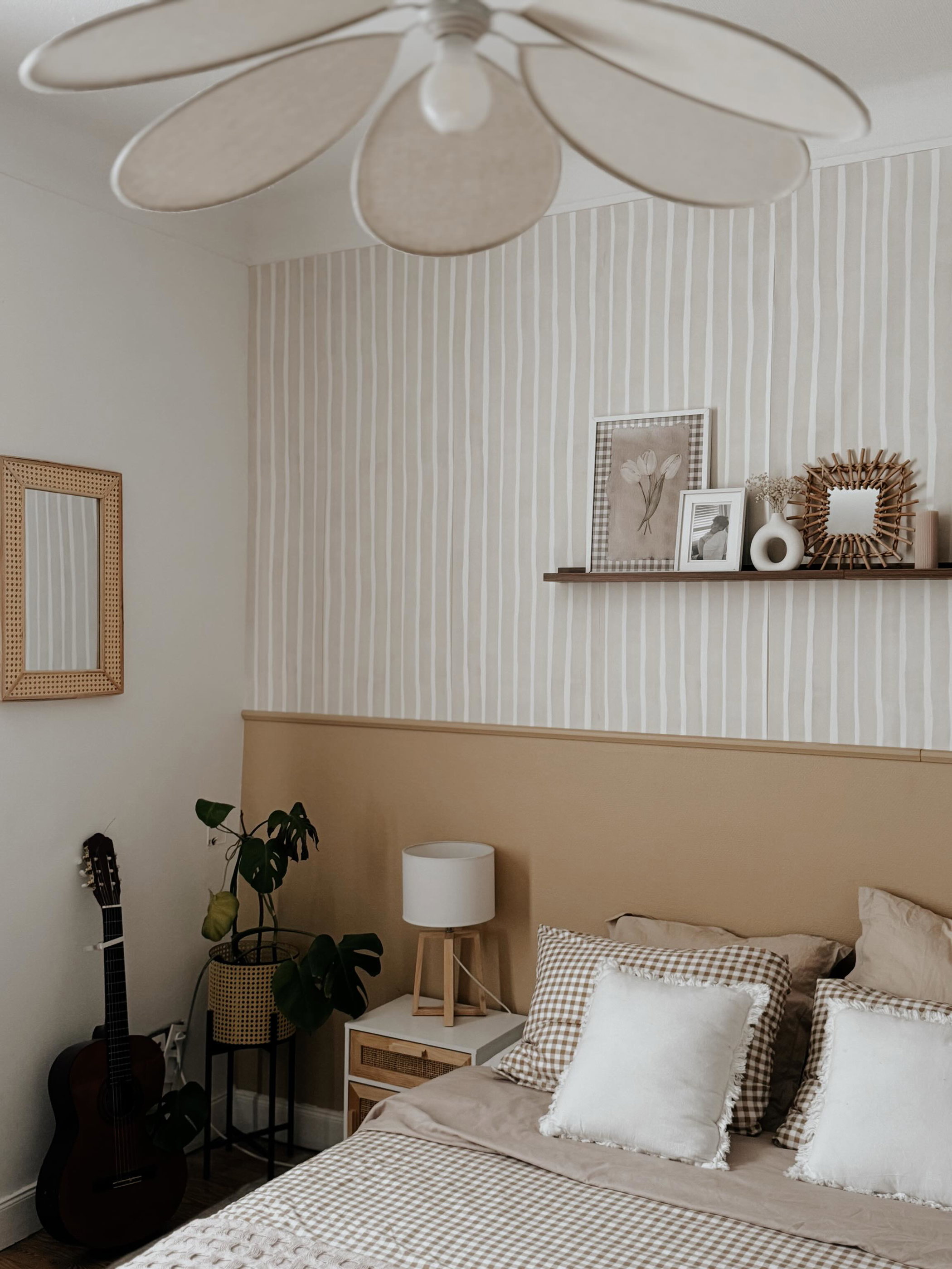 Ein neutral gestaltetes Schlafzimmer mit einem Bett, karierten und weißen Kissen, einem Nachttisch aus Holz und einer Gitarre. Beige gestreifte Tapete und ein schwebendes Regal mit Dekoration.