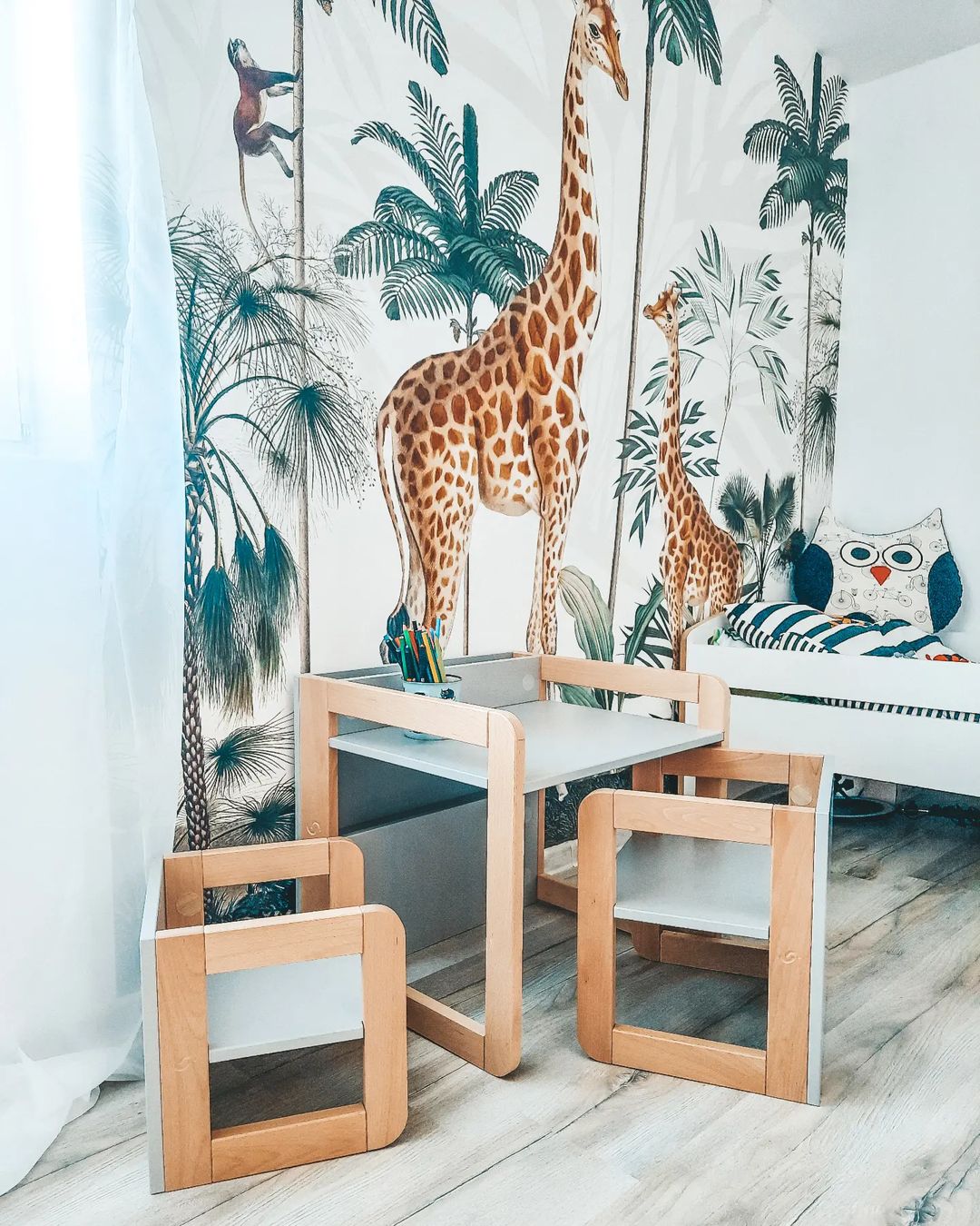 Chambre d'enfant avec papier peint jungle, table et chaises en bois, et un lit de jour avec un oreiller hibou.