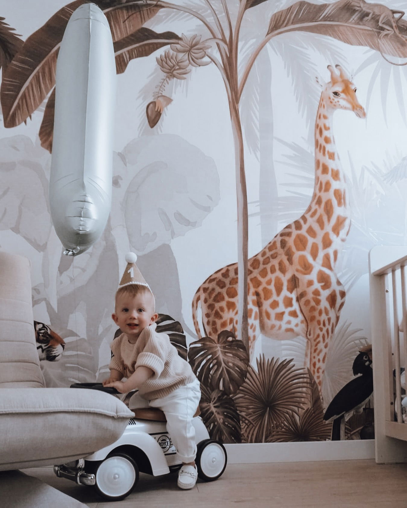 Een baby met een feesthoed zit op een speelgoedauto in een kamer met jungle-thema.