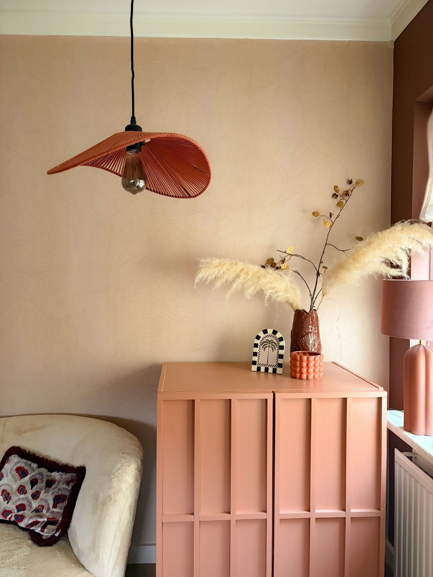 Salmon-colored cabinet with decorative items and pampas grass, under an orange pendant light.
