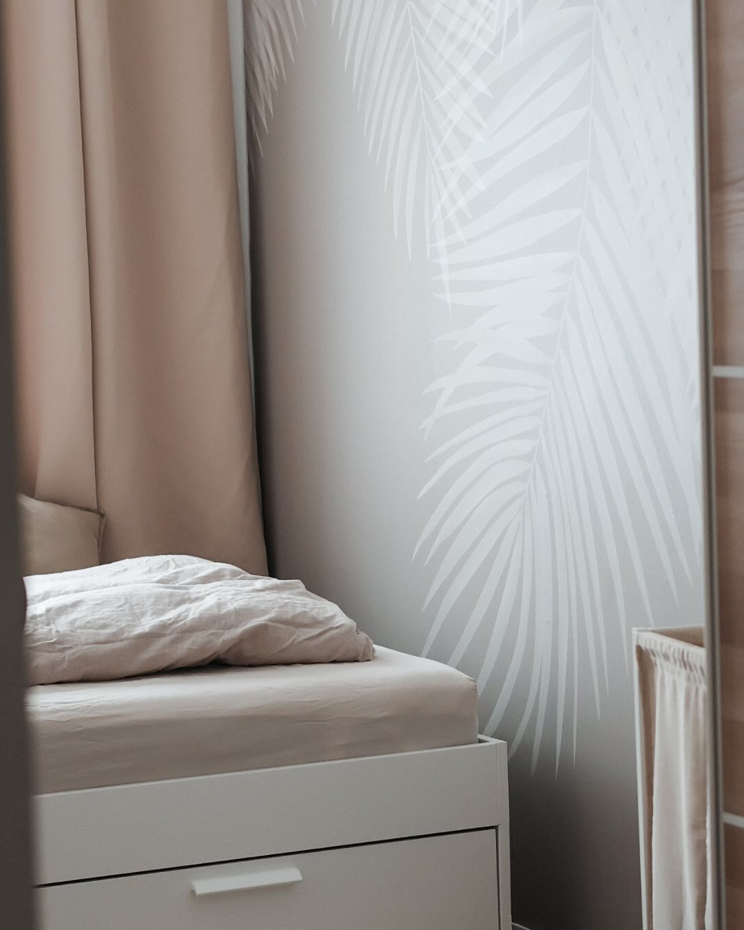 Esquina de dormitorio con estructura de cama blanca, ropa de cama beige y papel tapiz de hojas de palma.