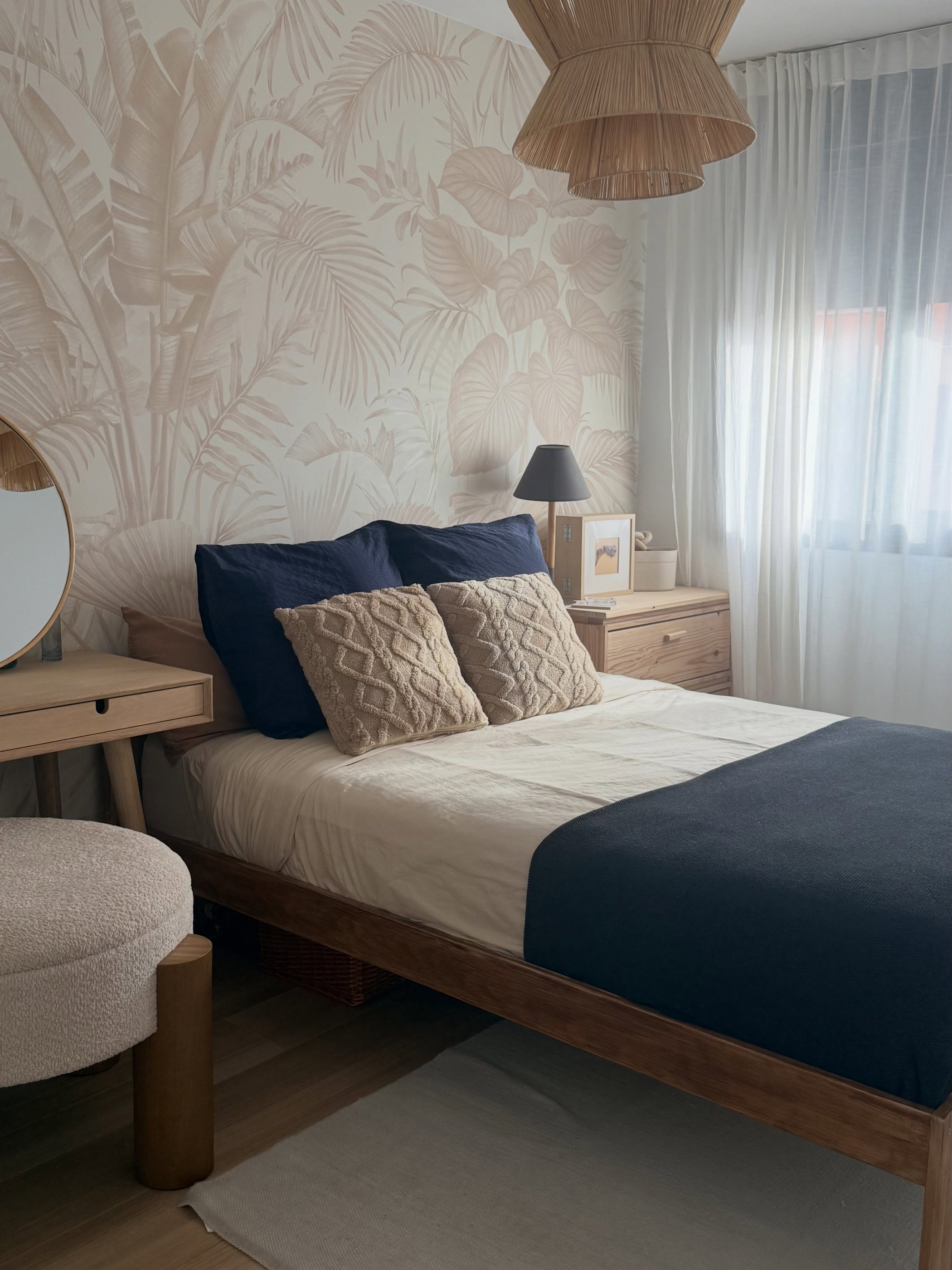 Bedroom with wooden bed, navy and beige bedding, beige tropical leaf wallpaper. Wood vanity with round mirror, fuzzy stool, woven pendant light.