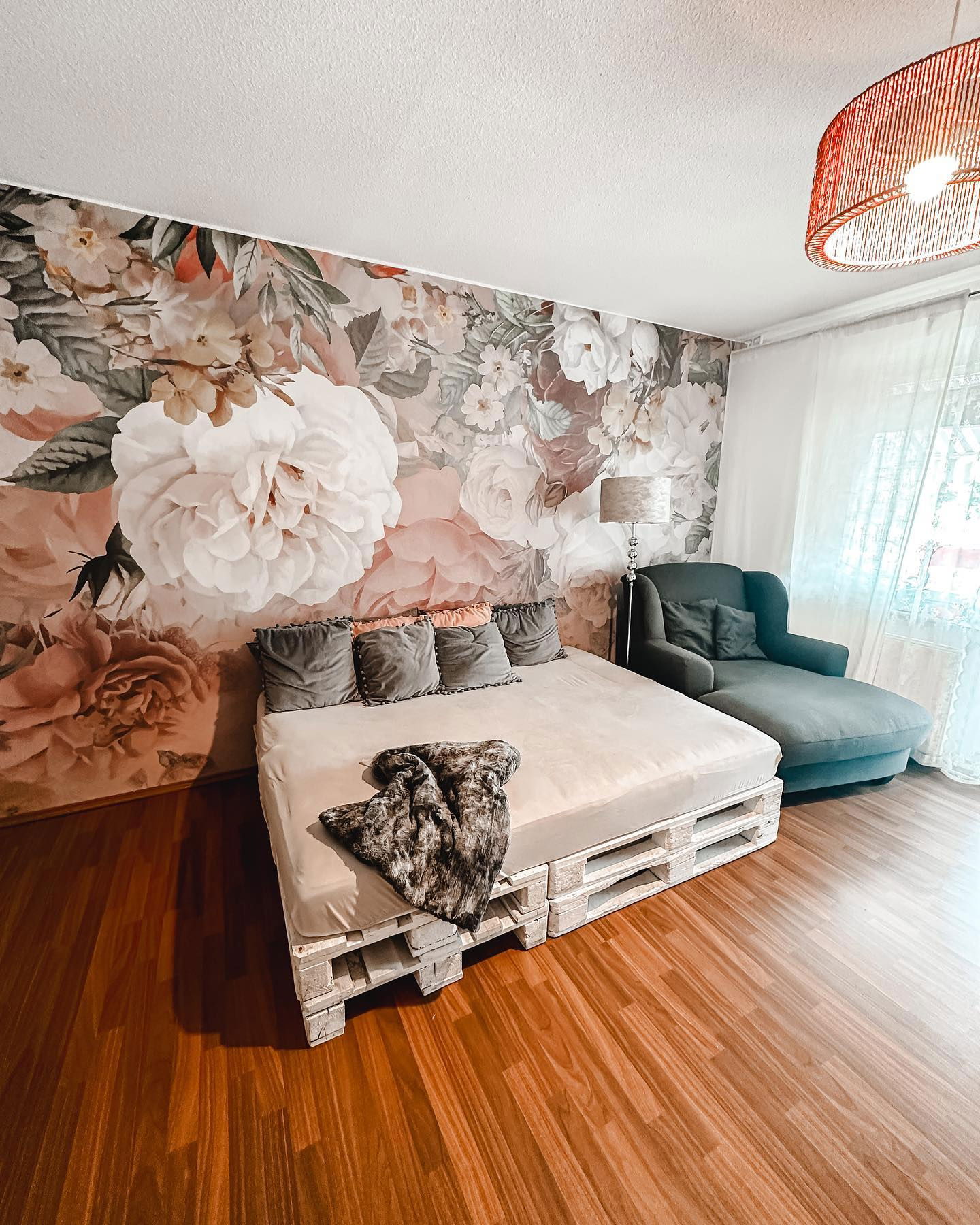 Dormitorio con papel tapiz floral, cama de paletas, almohadas grises y chaise lounge.