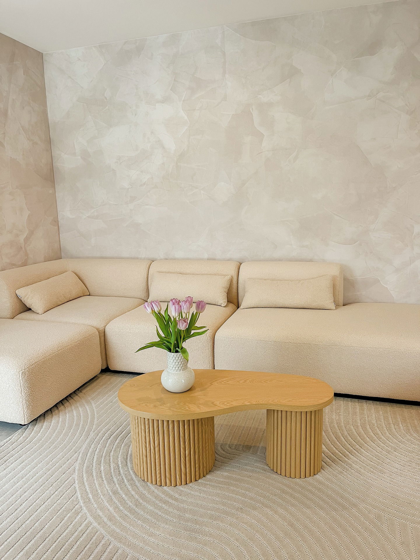 Beige sectional sofa with pillows, wooden coffee table with tulips in a vase, and a cream rug with wave pattern.