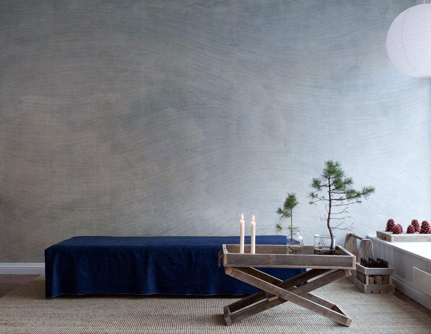 Minimalist interior with blue linen-covered bench, wooden tray table with candles and small pine trees.