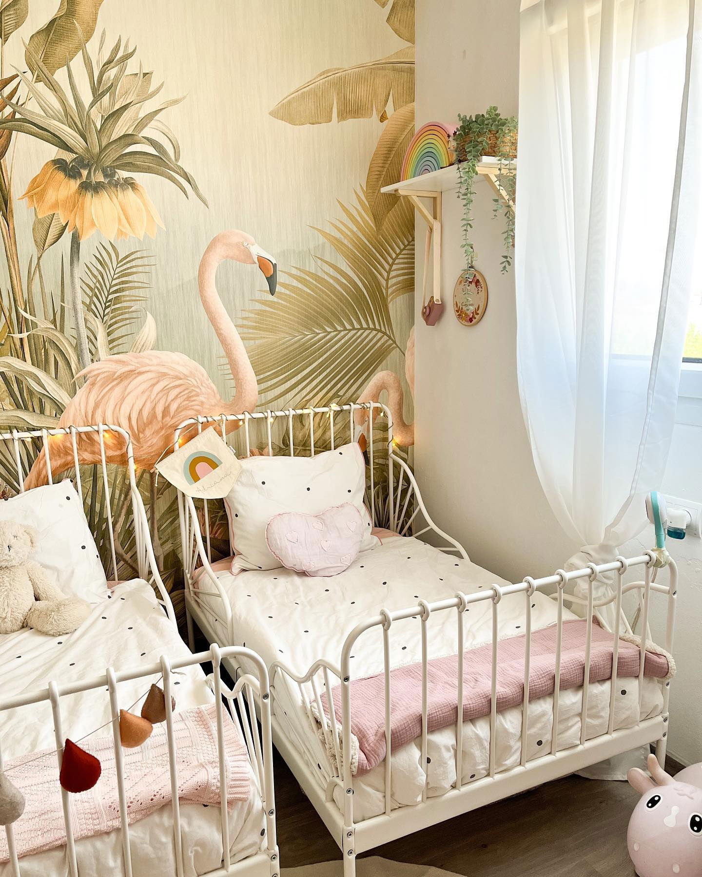 Kids' room with two white metal beds, flamingo wallpaper, and rainbow decor.