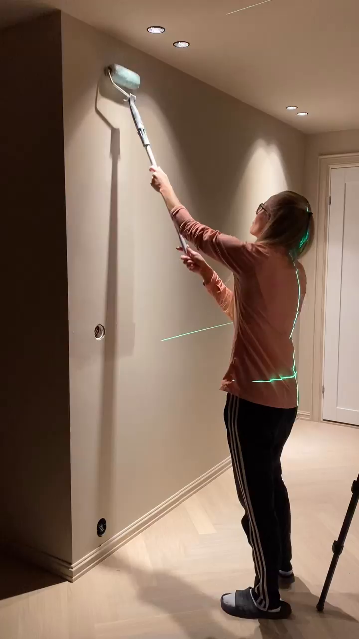 A woman uses a paint roller on a wall, illuminated by green laser lines.