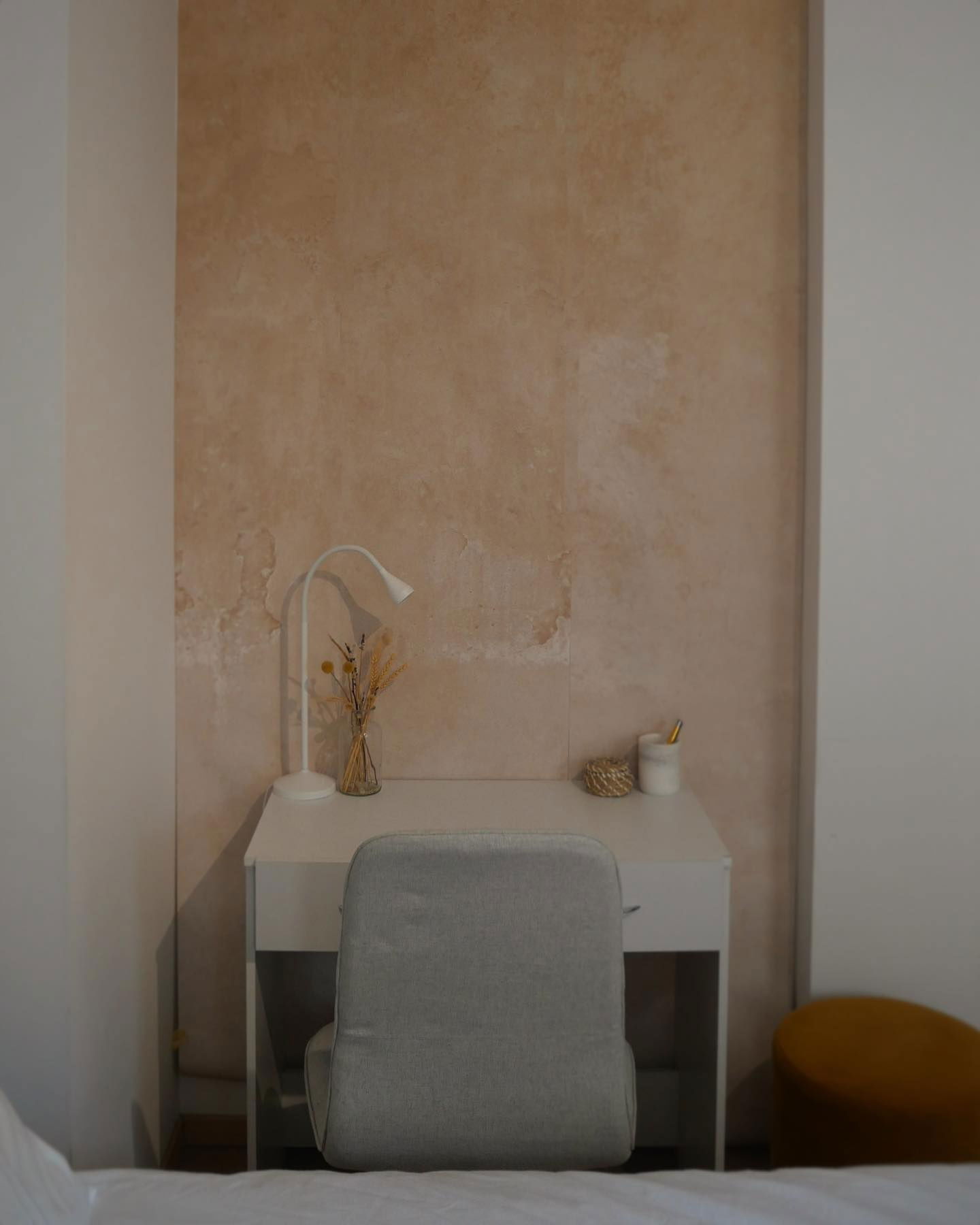Minimalistischer Arbeitsbereich mit weißem Schreibtisch, grauem Stuhl, Lampe und Trockenblumen vor einer strukturierten Wand.