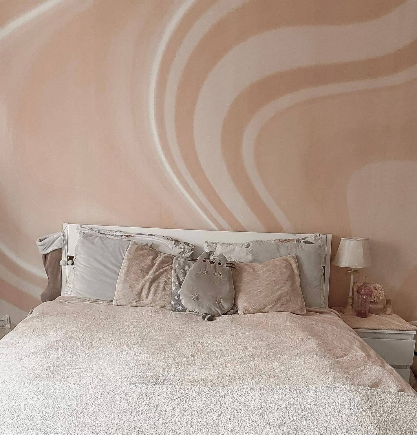 Soveværelse med pink vægkunst, hvid sengeramme og bløde puder. En Pusheen-plys sidder på sengen.