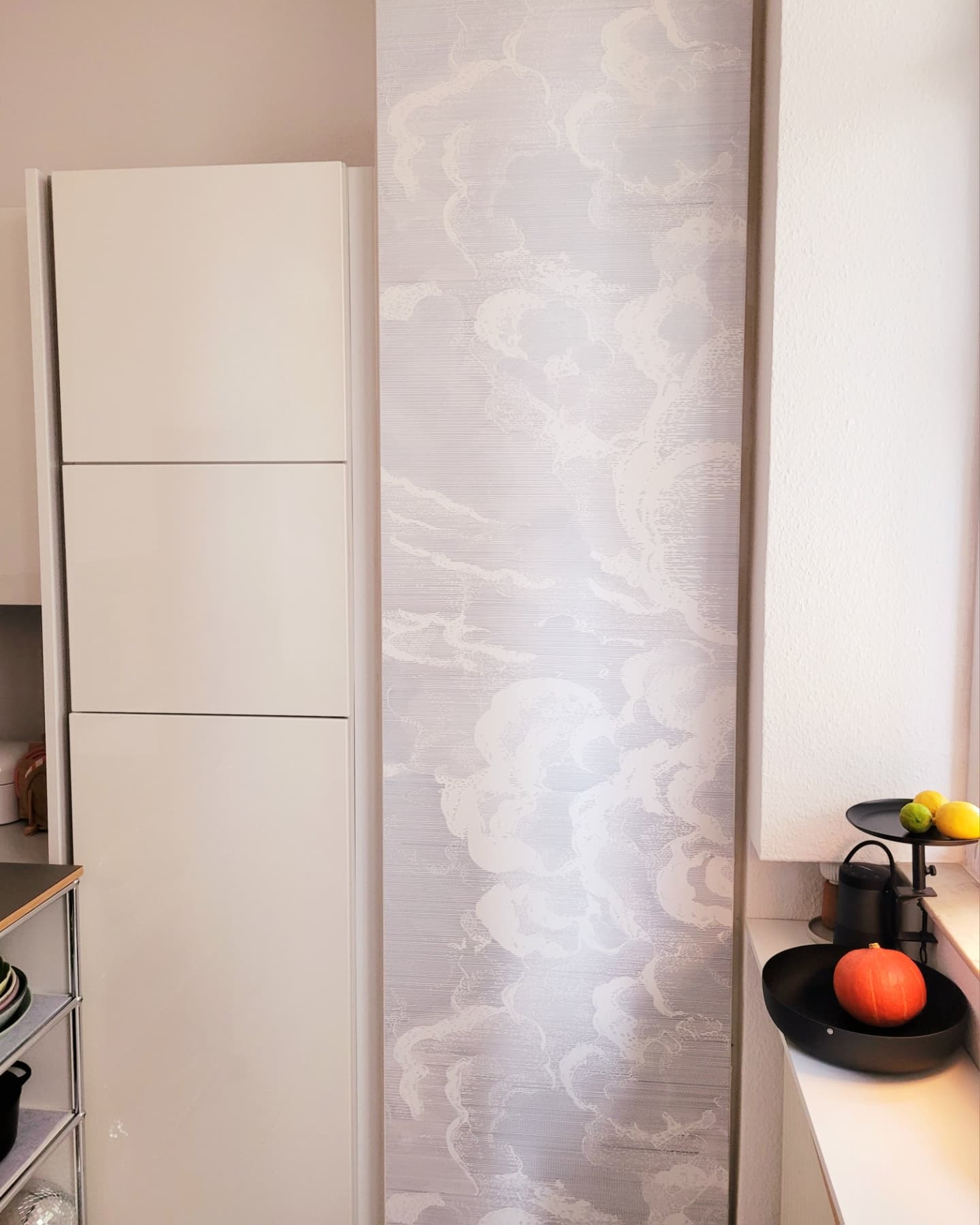 Innenansicht einer Küche mit weißen Schränken, Wolkenmuster-Tapete und einem Fensterbrett mit Dekorationsgegenständen.
