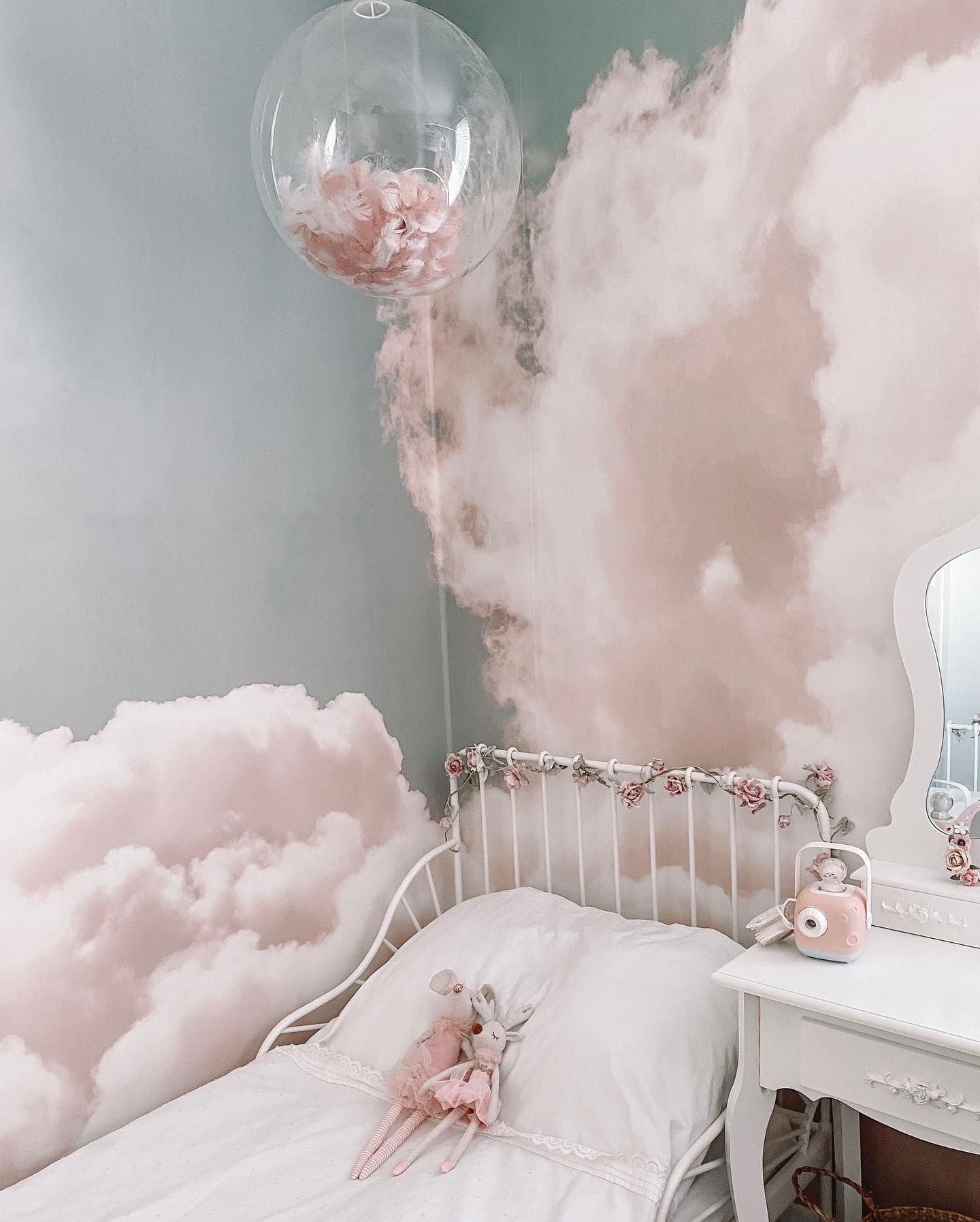 Child's bedroom with cloud wallpaper, white bed, and pink accents. A clear balloon filled with pink flowers hangs above.