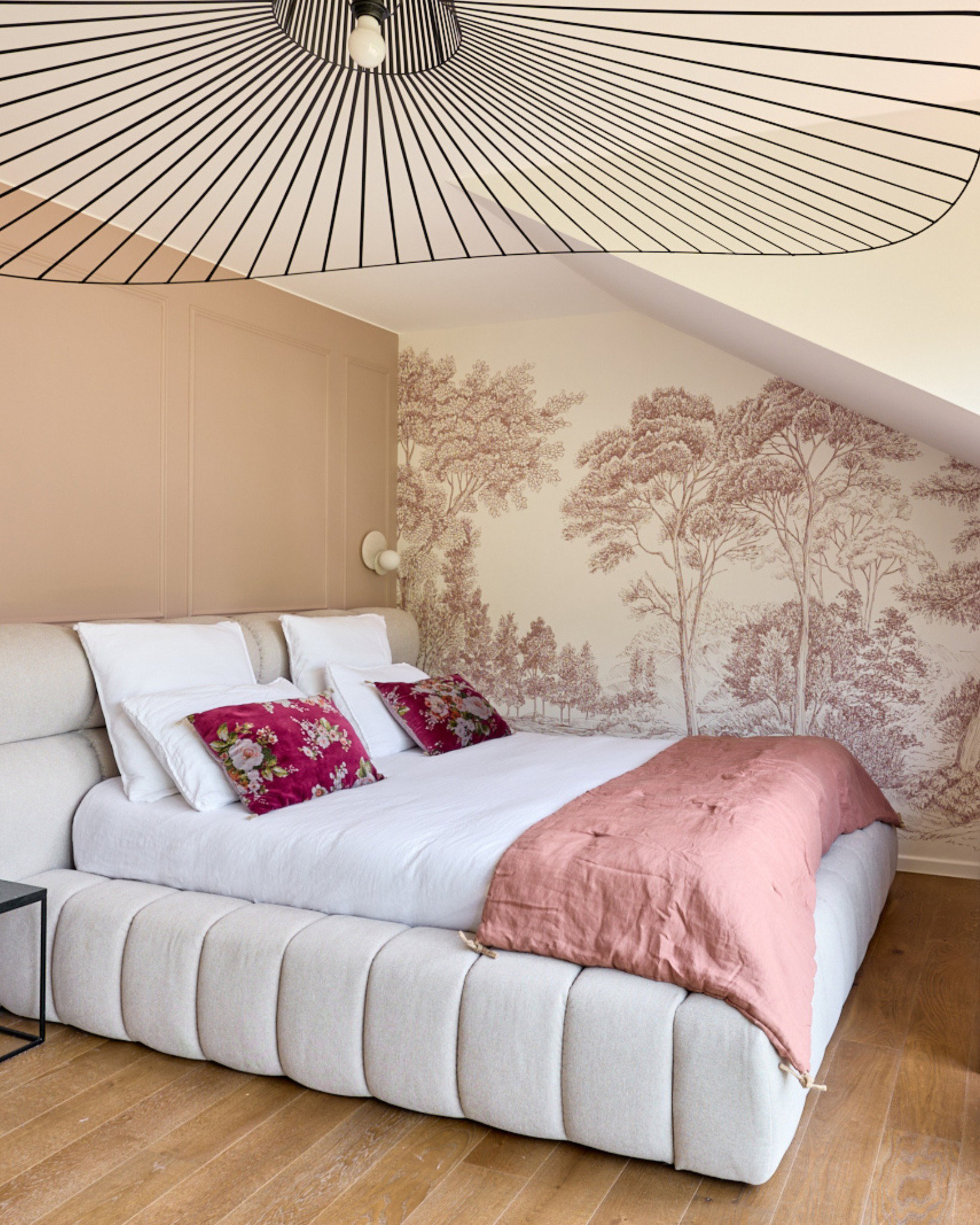 Ett rymligt sovrum har en ljusbeige stoppad säng med vita sängkläder, två vinröda blommigade kuddar och en rosa pläd. En vägg har beige tapet med ett vinrött träd-mönster. En svartvit fläktliknande pendellampa hänger från taket.