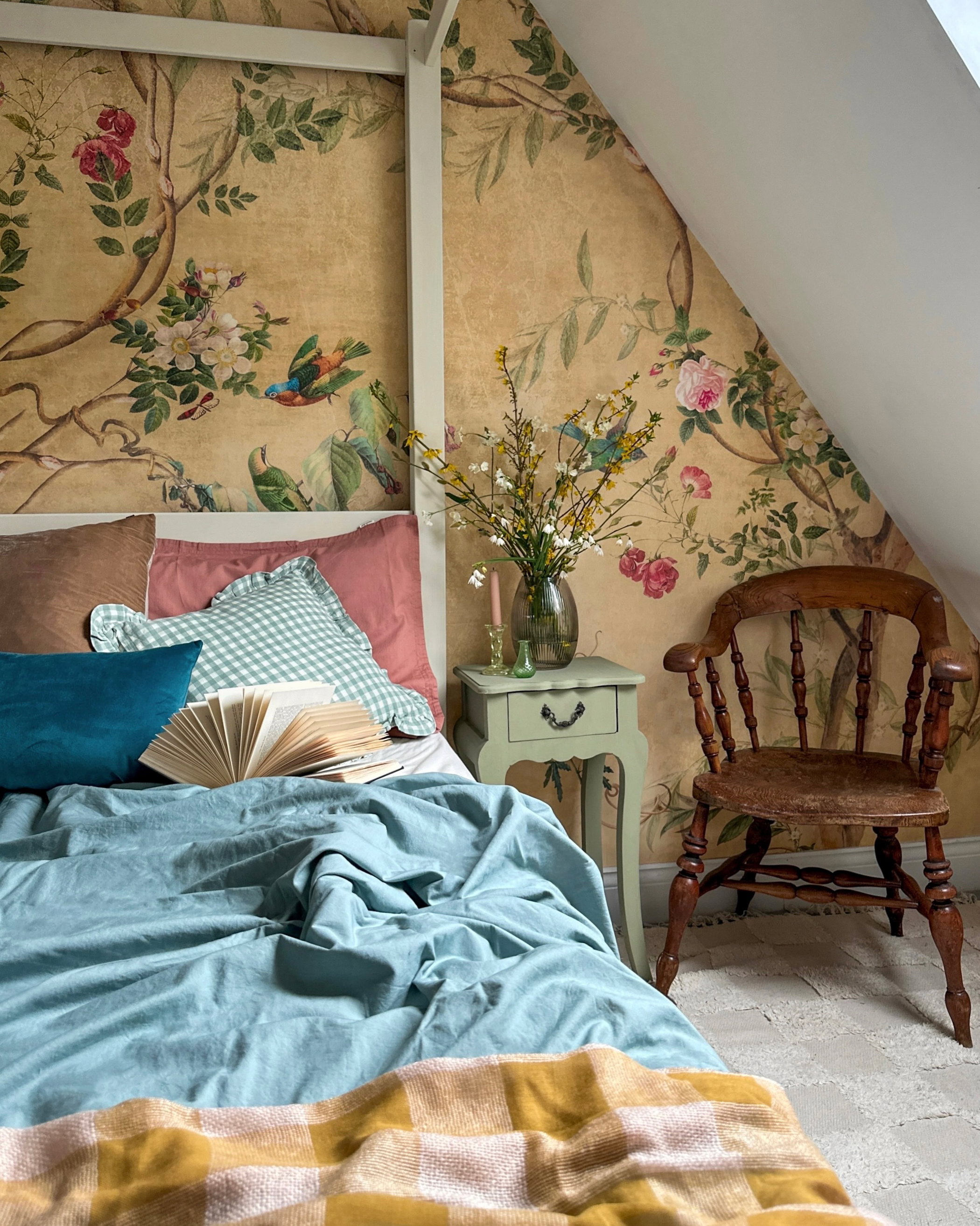 Soveværelse med en seng, lyseblåt dynebetræk og et gult plaid-tæppe. Beige vægge har et levende blomster- og fugletapet. Et grønt natbord indeholder blomster.
