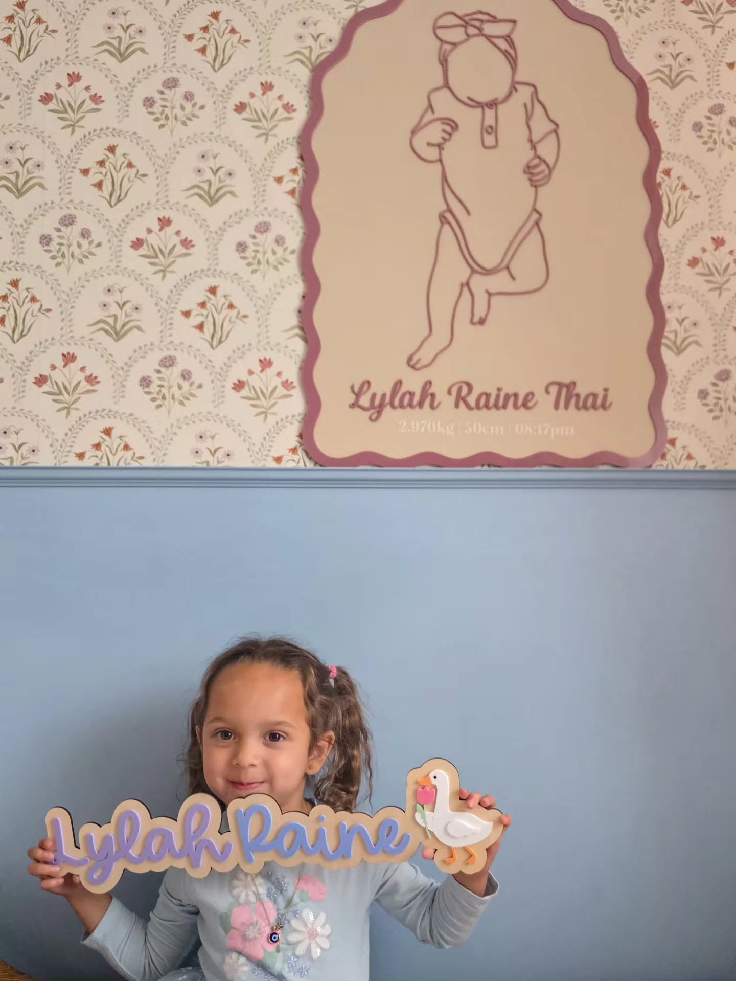 Joven sonriente sostiene un cartel de madera con "Lylah Raine" escrito en morado, junto a un recorte de ganso blanco. Detrás de ella, un cartel de madera más grande muestra la silueta de un bebé y "Lylah Raine Thai" sobre un papel tapiz floral.