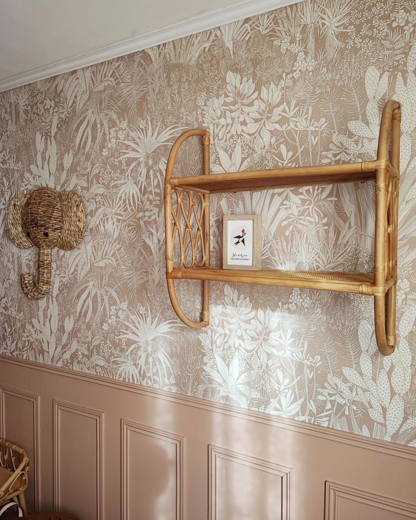 Pared interior con papel tapiz floral, revestimiento de madera rosa, cabeza de elefante tejida y estante de ratán con arte enmarcado.