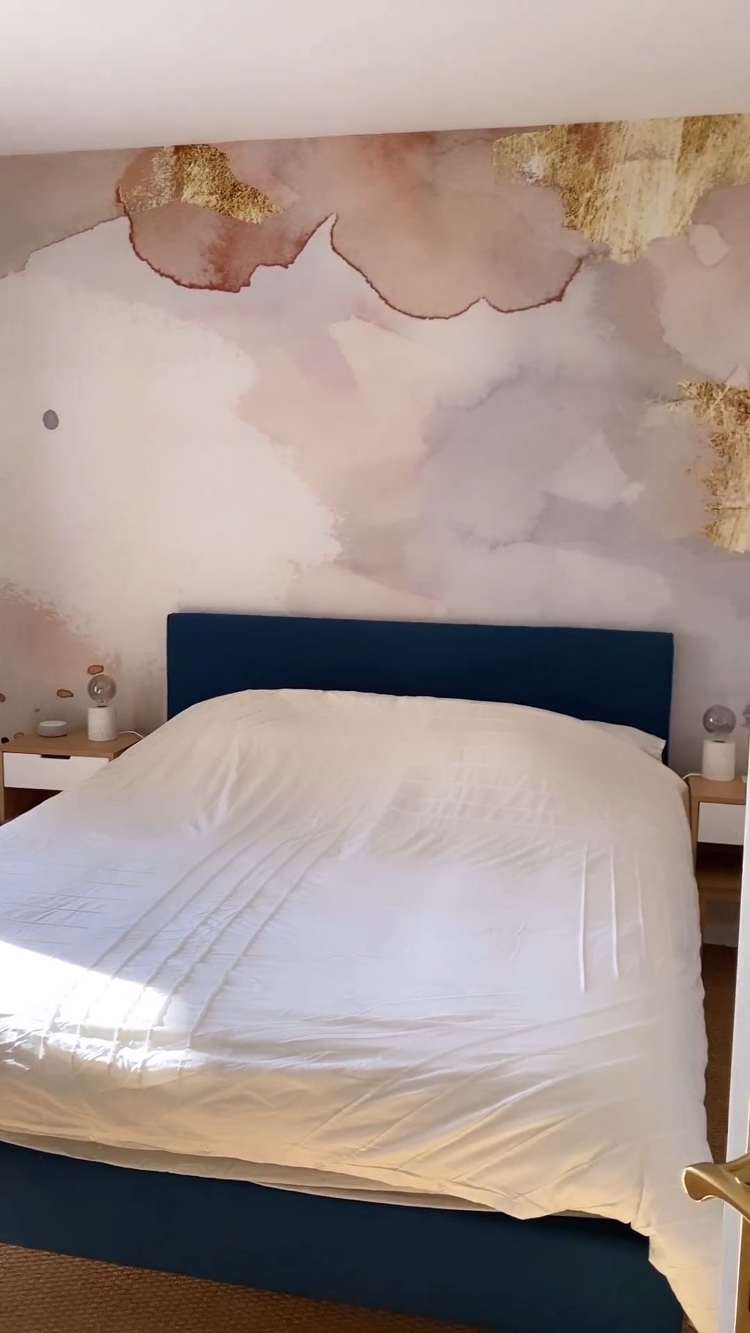 Bedroom with blue headboard, leaf-patterned duvet, striped wallpaper, and white bedside table.