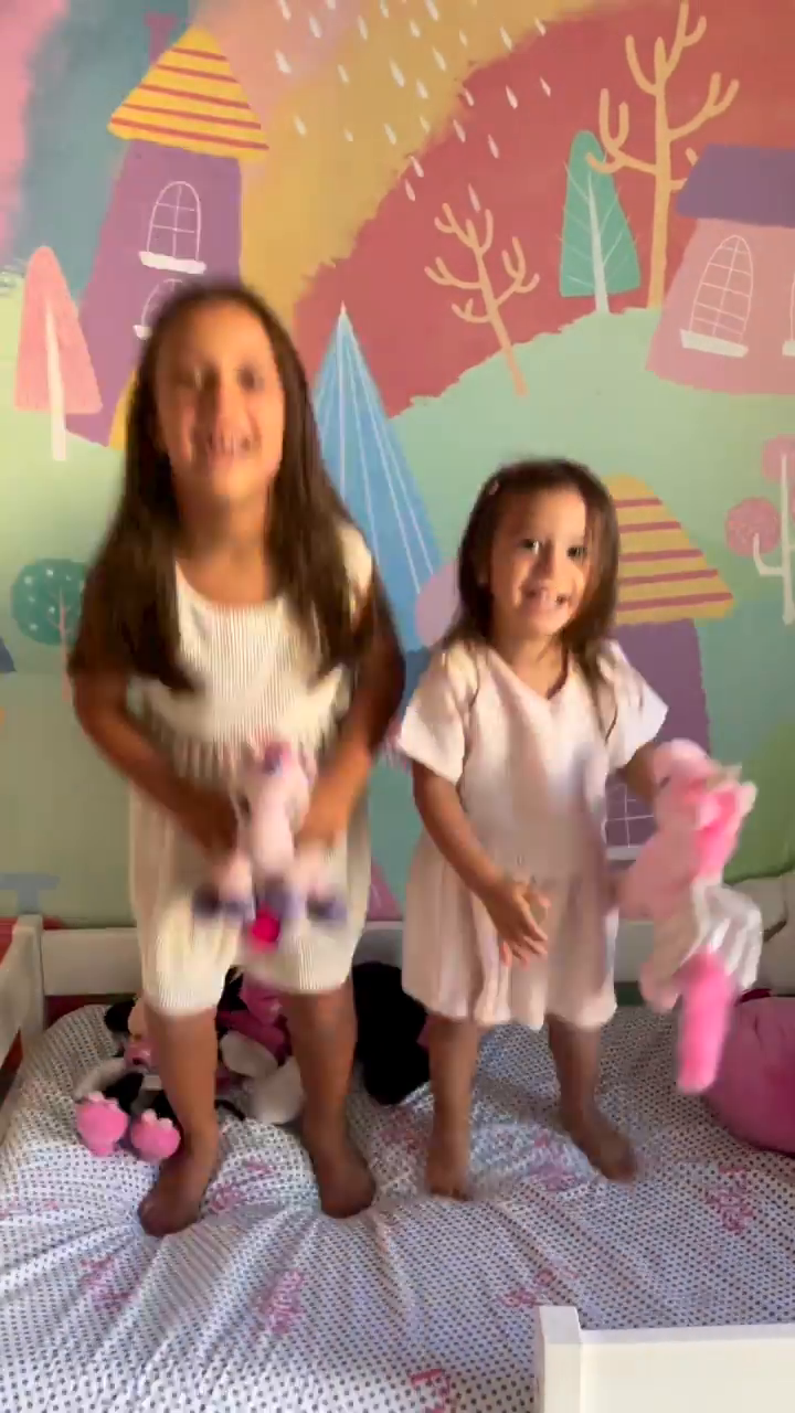 Two young girls jumping on a bed with stuffed animals and a colorful mural.