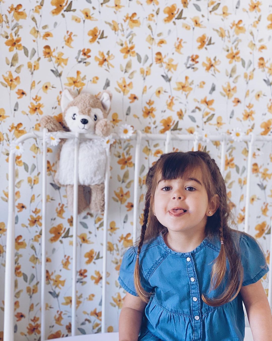 A young girl with pigtails sticks out her tongue in front of a floral wallpaper. A stuffed animal hangs on the crib behind her.