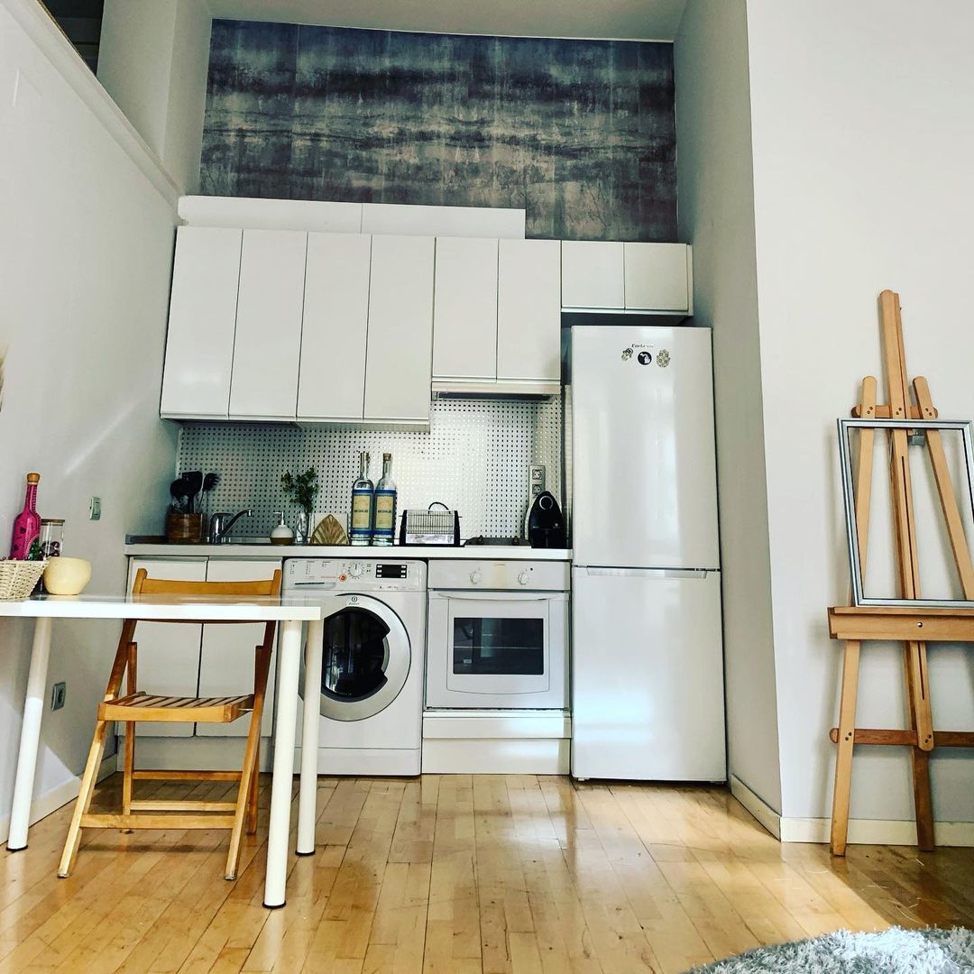 Compact kitchen with white cabinets, appliances, and a small dining table with a wooden chair. An easel stands nearby.
