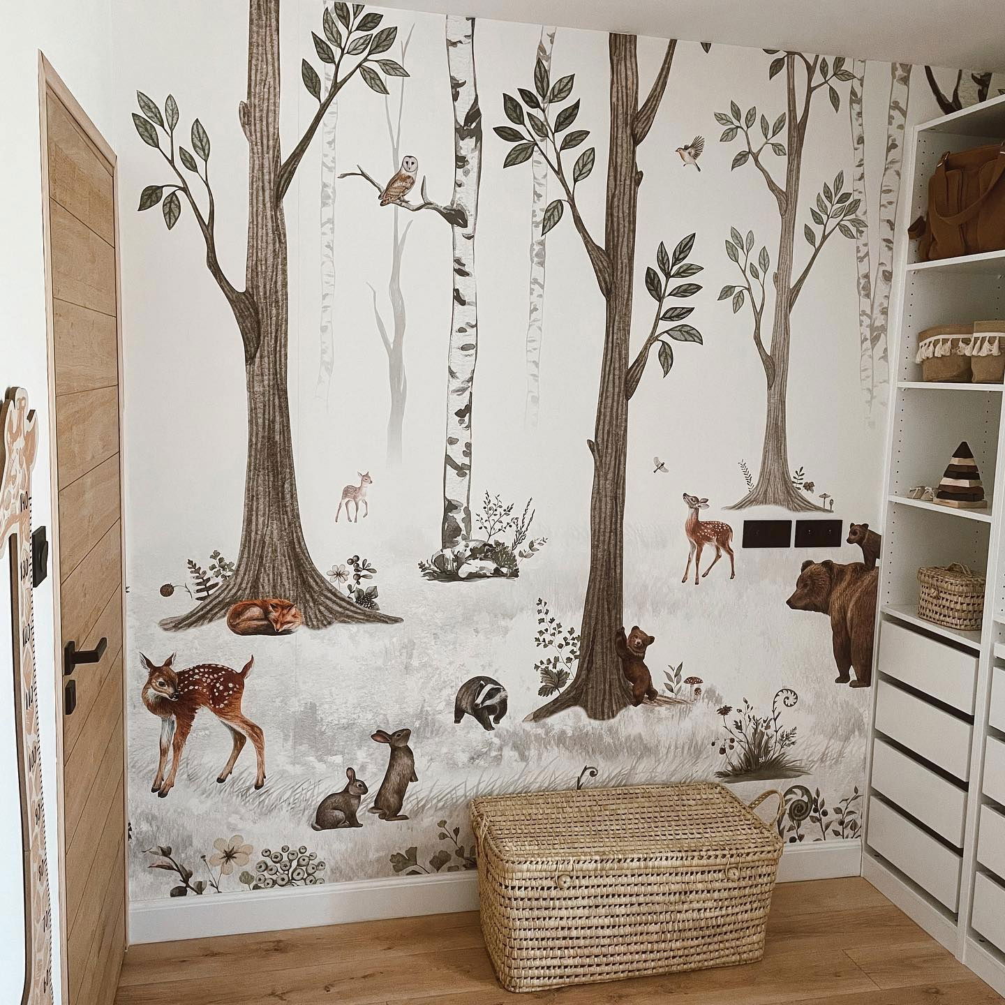 Nursery with woodland-themed wallpaper featuring trees, deer, bears, rabbits, and an owl. A woven storage chest sits on the wooden floor.