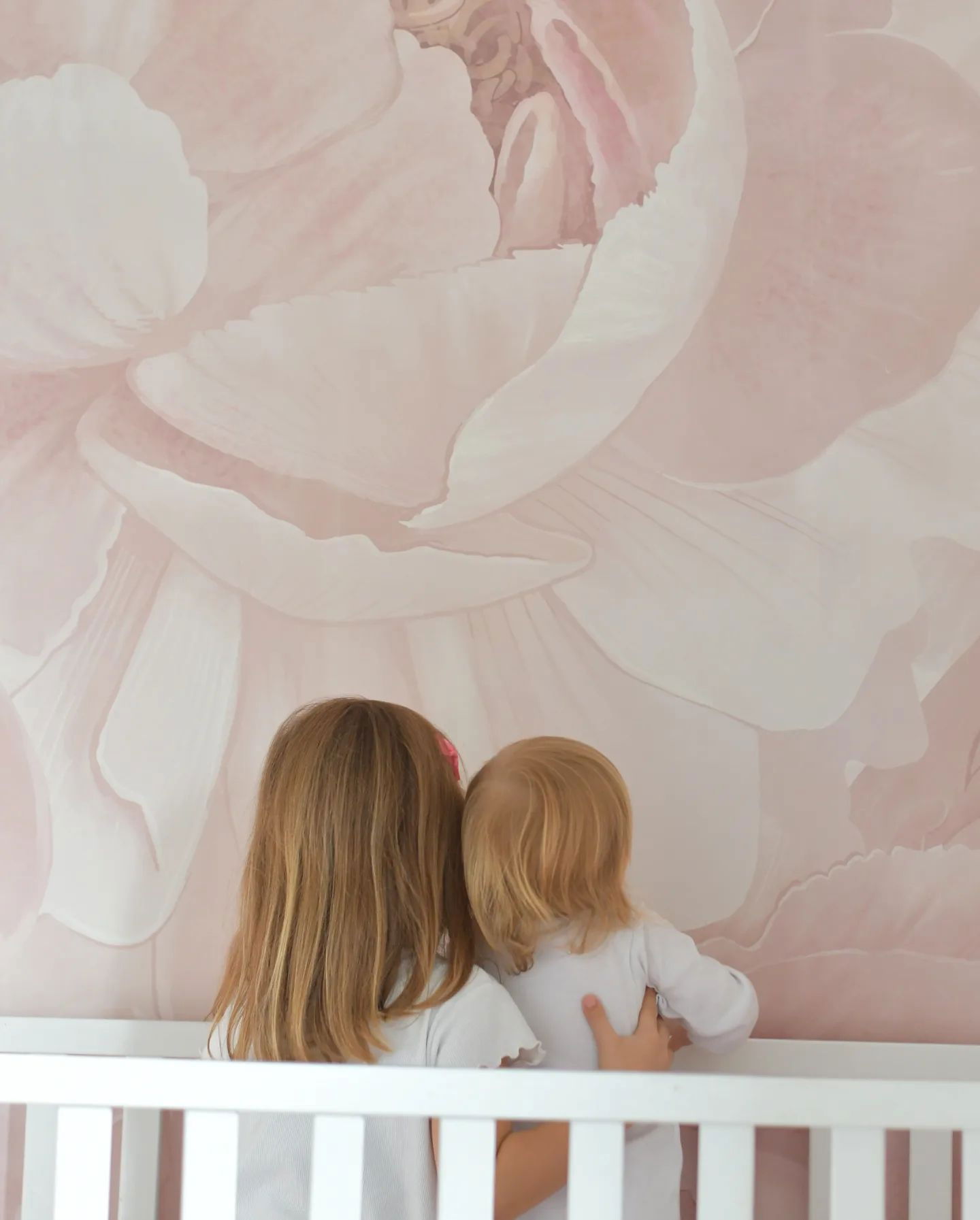 Due bambini piccoli sono in piedi in una culla bianca, fissando un grande murale da parete floreale rosa.