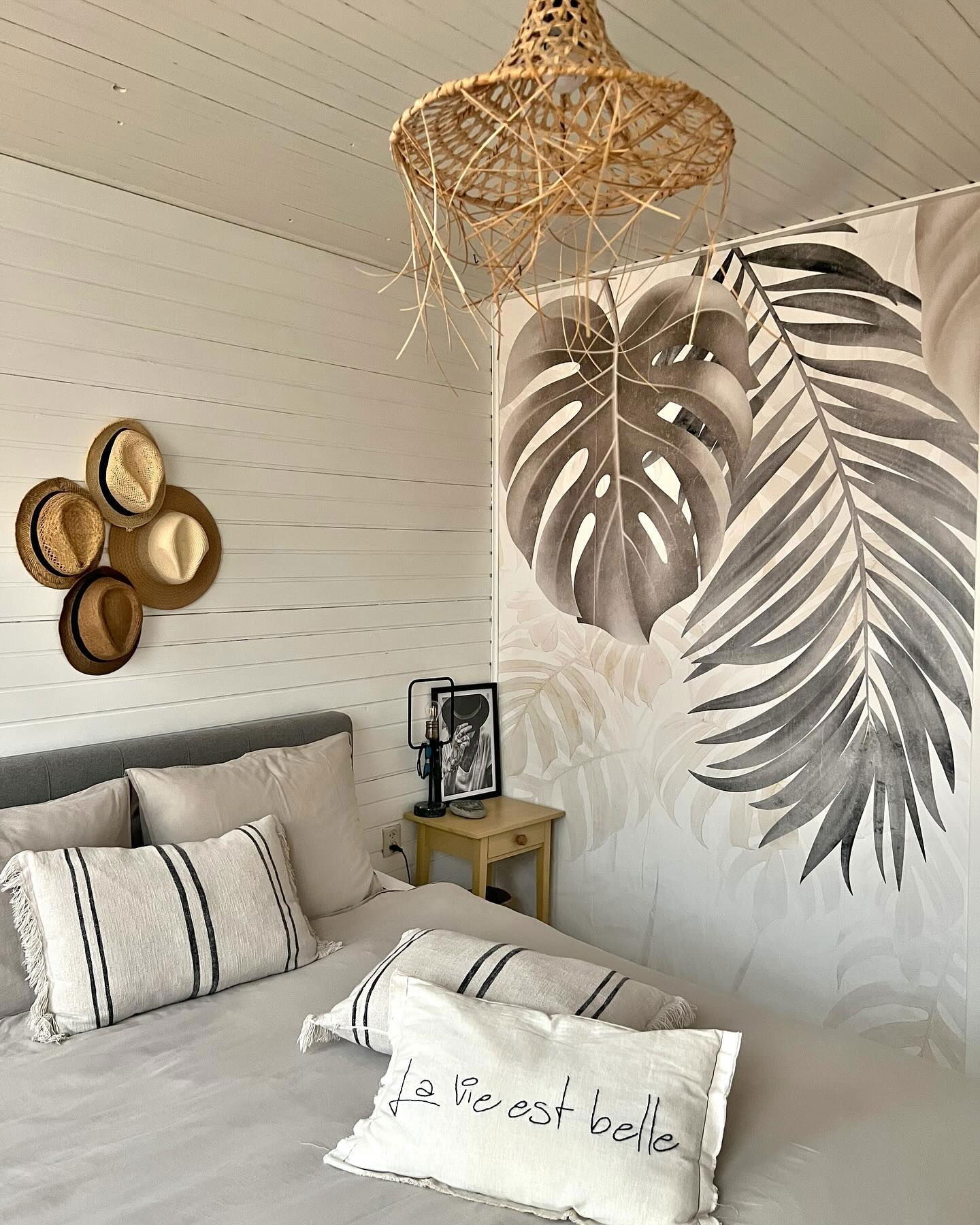 Bedroom with gray bedding, straw light fixture, and tropical leaf wallpaper. Hats hang on the wall. A pillow reads "La vie est belle."