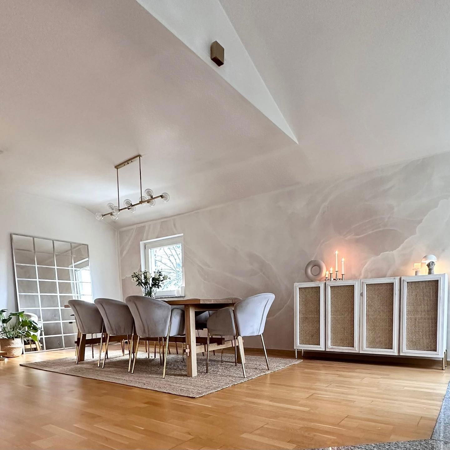 Sala da pranzo con pavimenti in legno, tavolo e sedie in velluto grigio. Un mobile bianco con ante in canna di Vienna è appoggiato a una parete con effetto marmo.