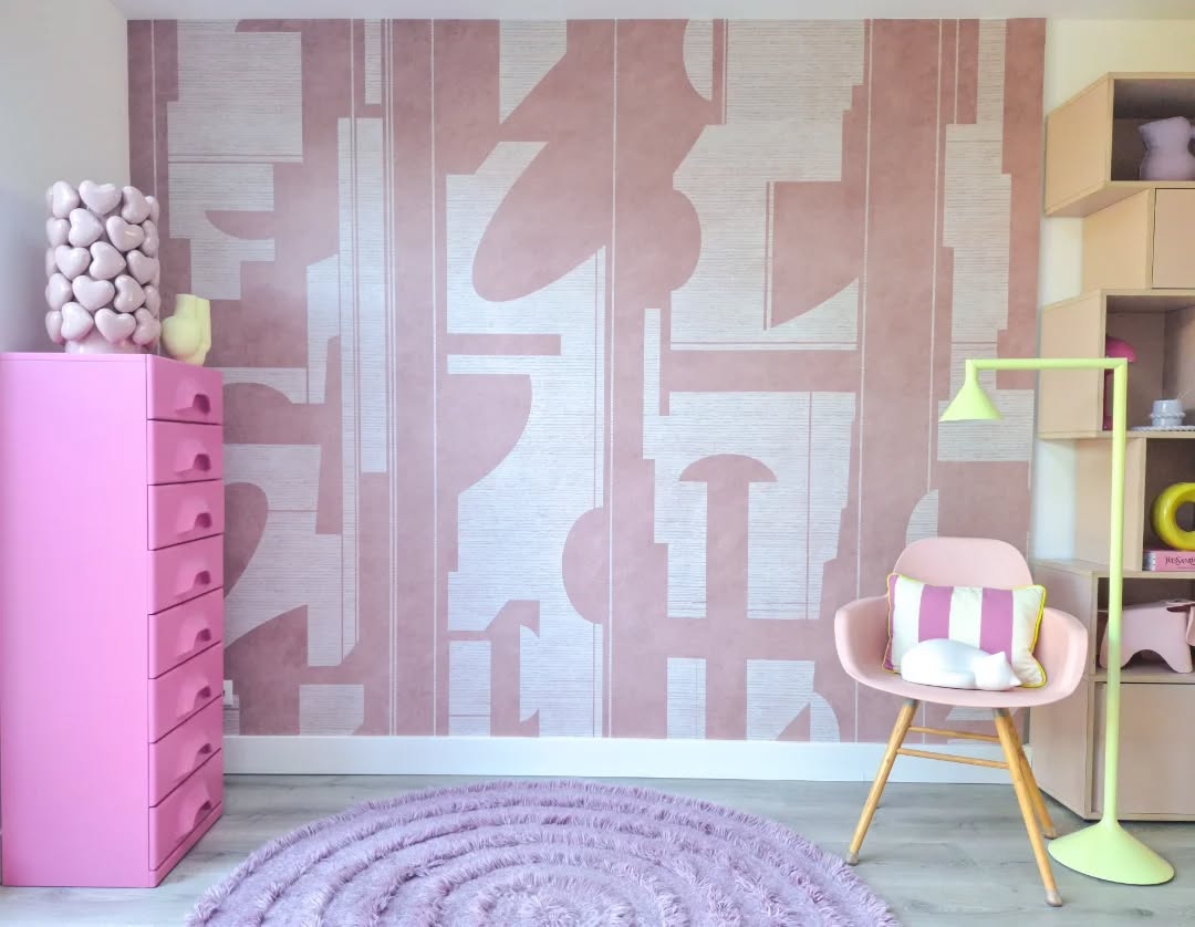 Pink dresser and chair in a room with patterned wallpaper and a yellow lamp.
