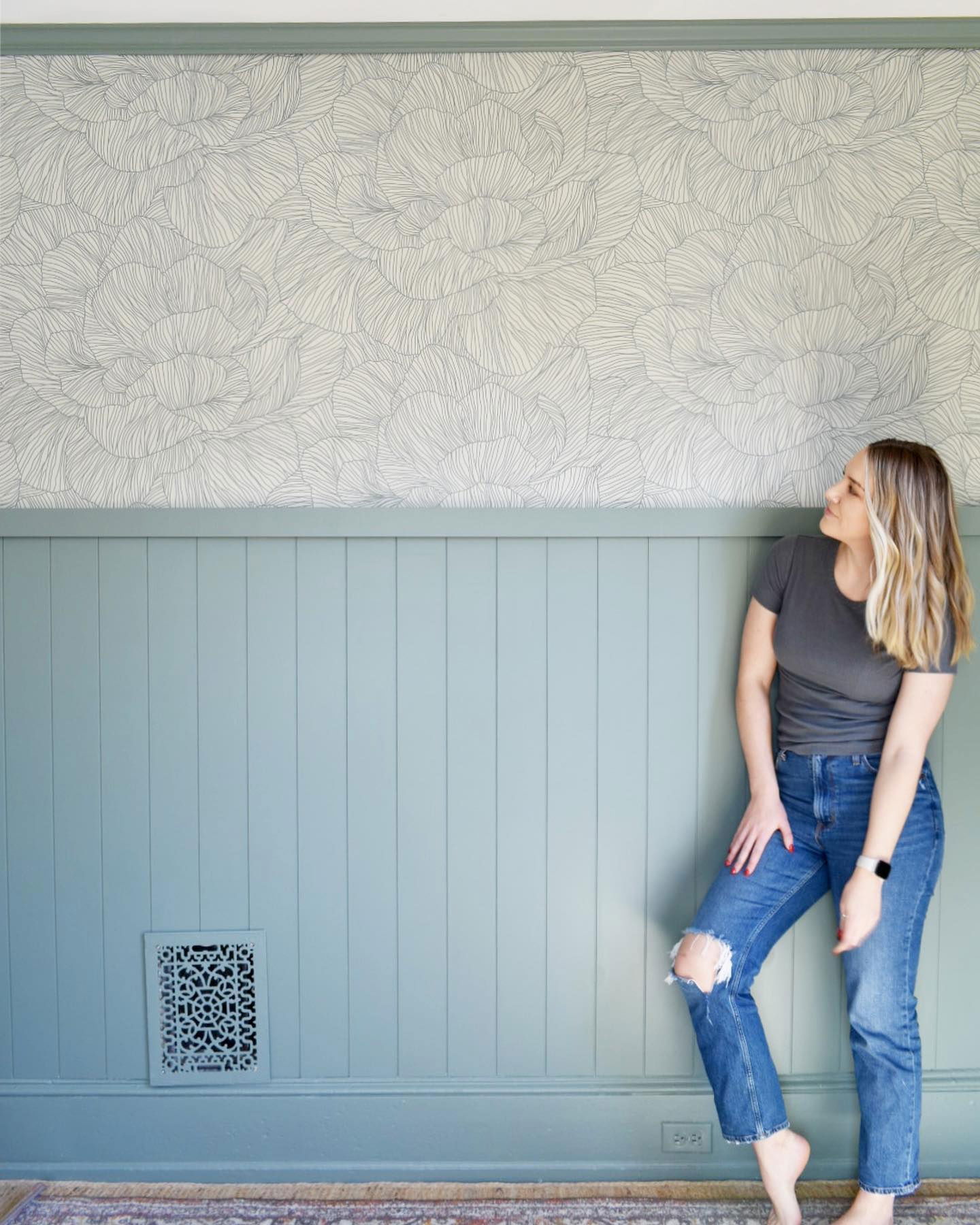 Donna che ammira una carta da parati floreale sopra una boiserie blu. Indossa jeans e una t-shirt grigia.