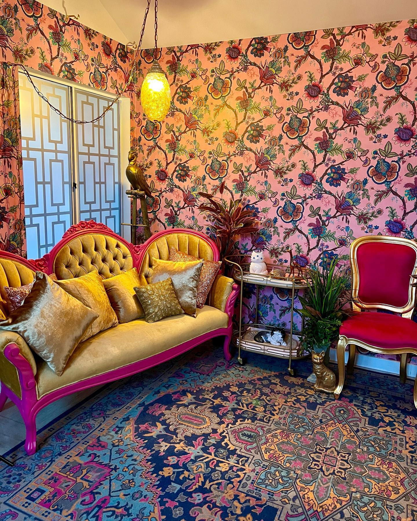 Eclectic living room with pink floral wallpaper, gold sofa with pink trim, and patterned rug.