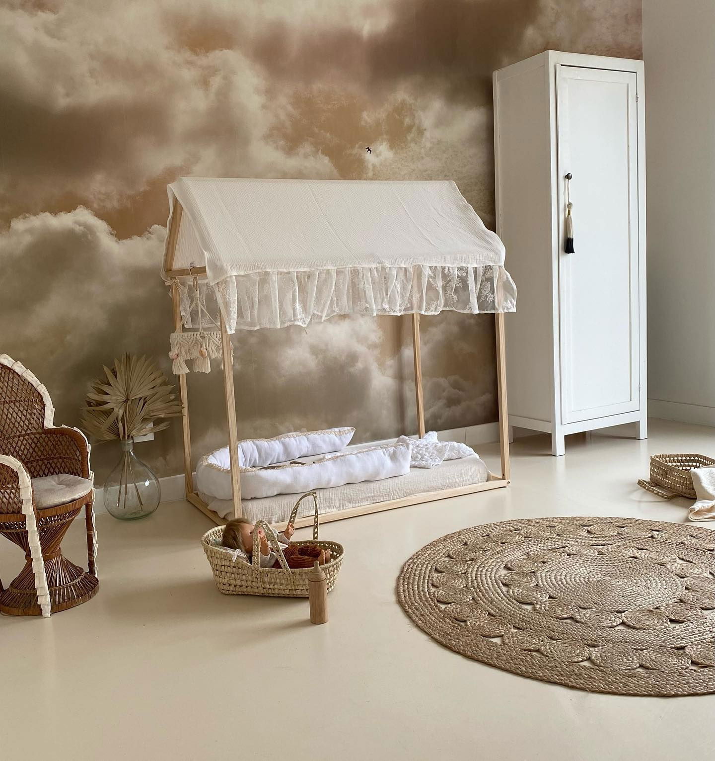 Nursery with white canopy bed, wicker chair, and round jute rug. Doll in woven basket with wooden bottle. Cloud wallpaper.