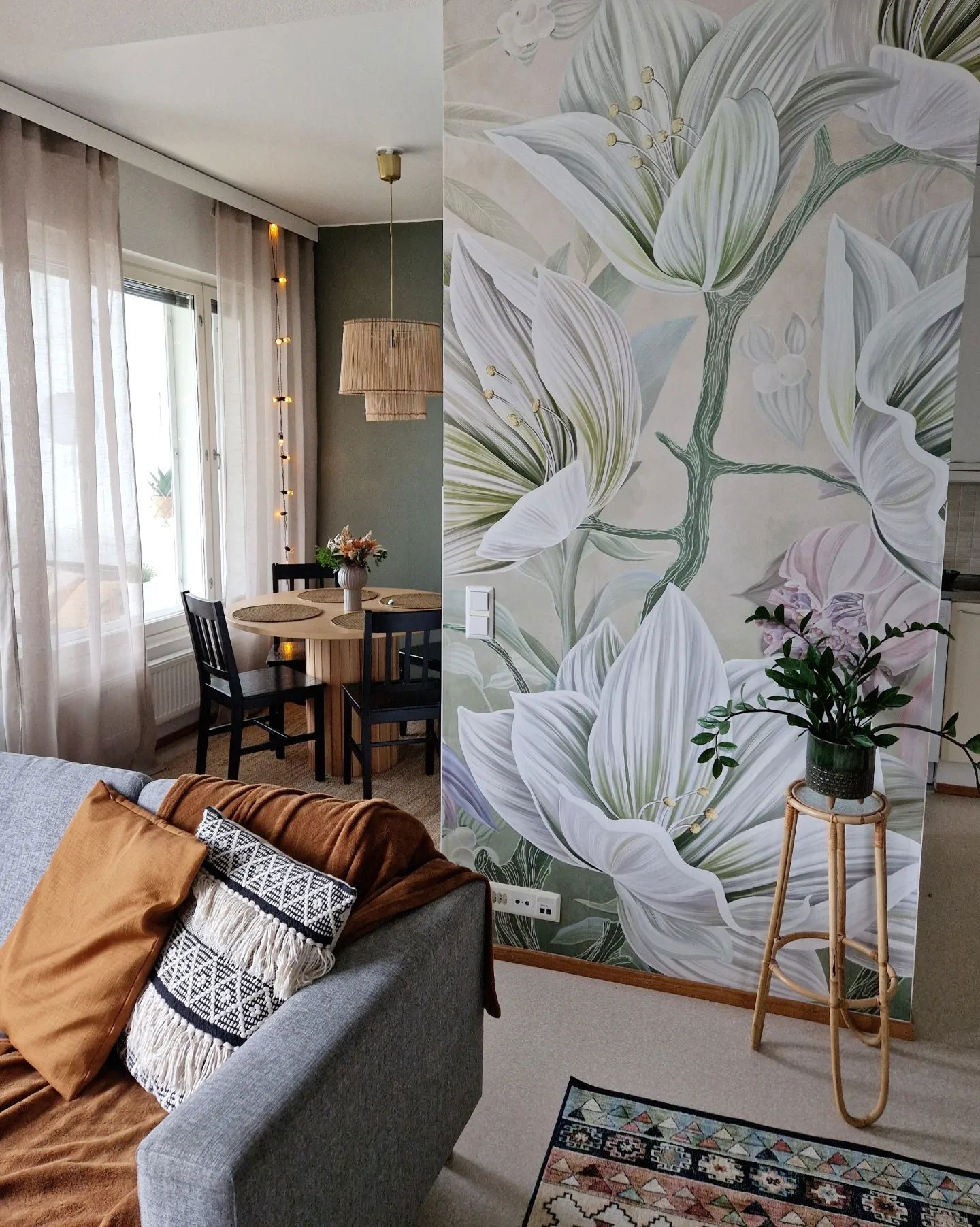 Sala de estar con mural floral, sofá gris con almohadas y mesa de comedor.