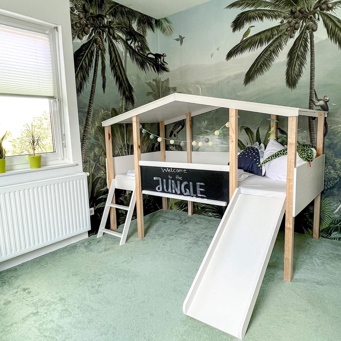 Child's jungle-themed bedroom with a white house frame bed, slide, and palm tree wallpaper.