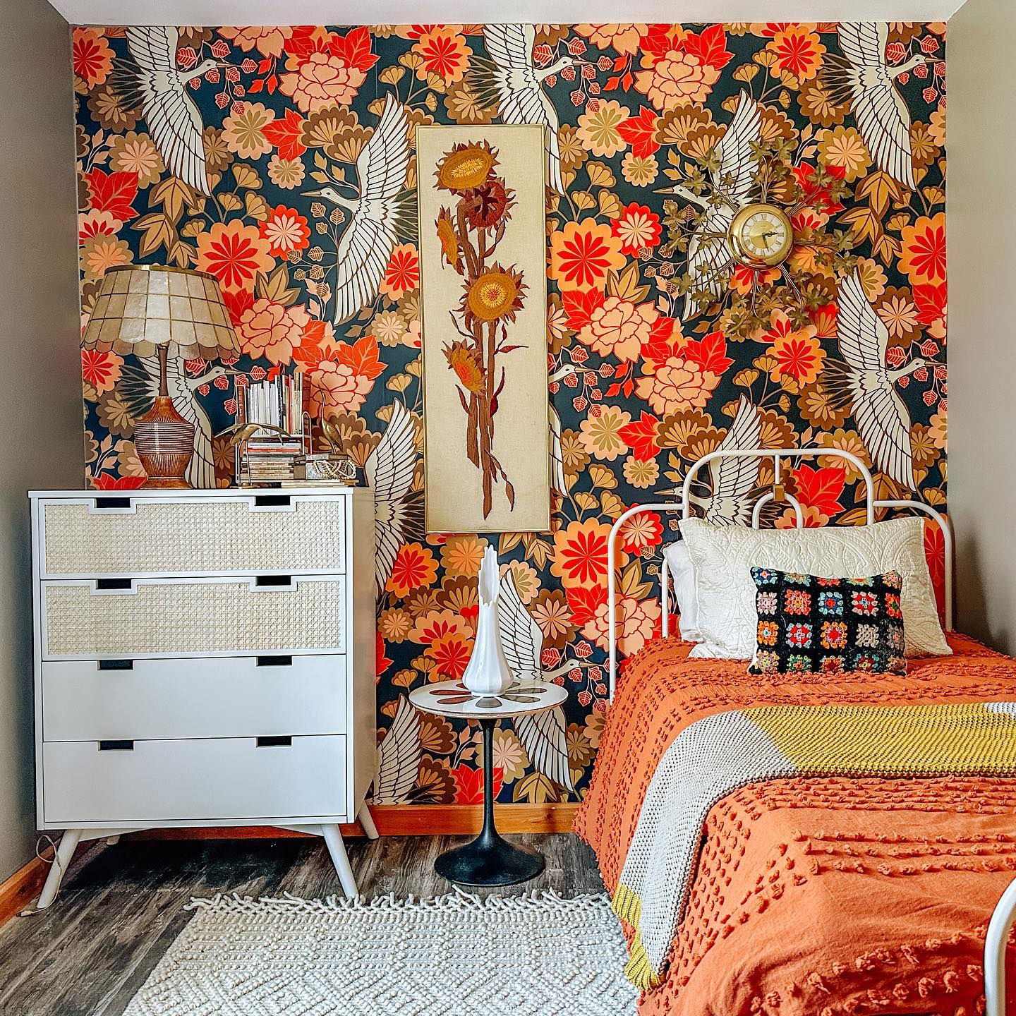 Cozy bedroom with floral wallpaper, white metal bed, and orange textured blanket. White dresser and small table.