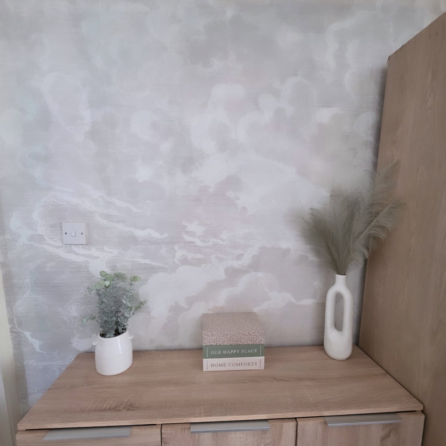 Interior with light wood dresser, decorative books, plant, and vase with pampas grass against a cloud-patterned wall.