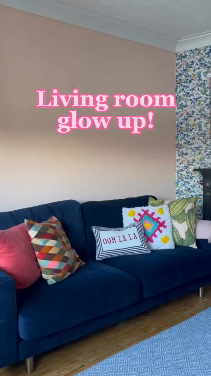 Living room with blue velvet sofa and colorful pillows. Wall is half pink paint, half floral wallpaper.